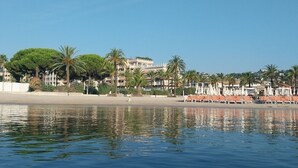 A calm waterfront with a beach, palm trees, and a building with balconies. - Hôtel Cecil (Antibes)