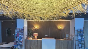 A reception desk with a wooden finish, a hanging woven canopy, and a wall-mounted lamp. - Hôtel Cecil (Antibes)