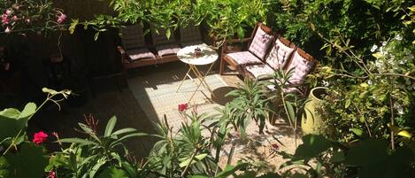Un patio soleado con zona de estar acolchada, rodeado de abundante follaje y plantas en macetas.