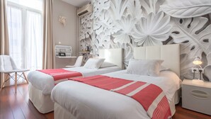A hotel room with two beds, a large floral wall mural, a desk with a chair, and a window with curtains. - Hôtel Cecil (Antibes)
