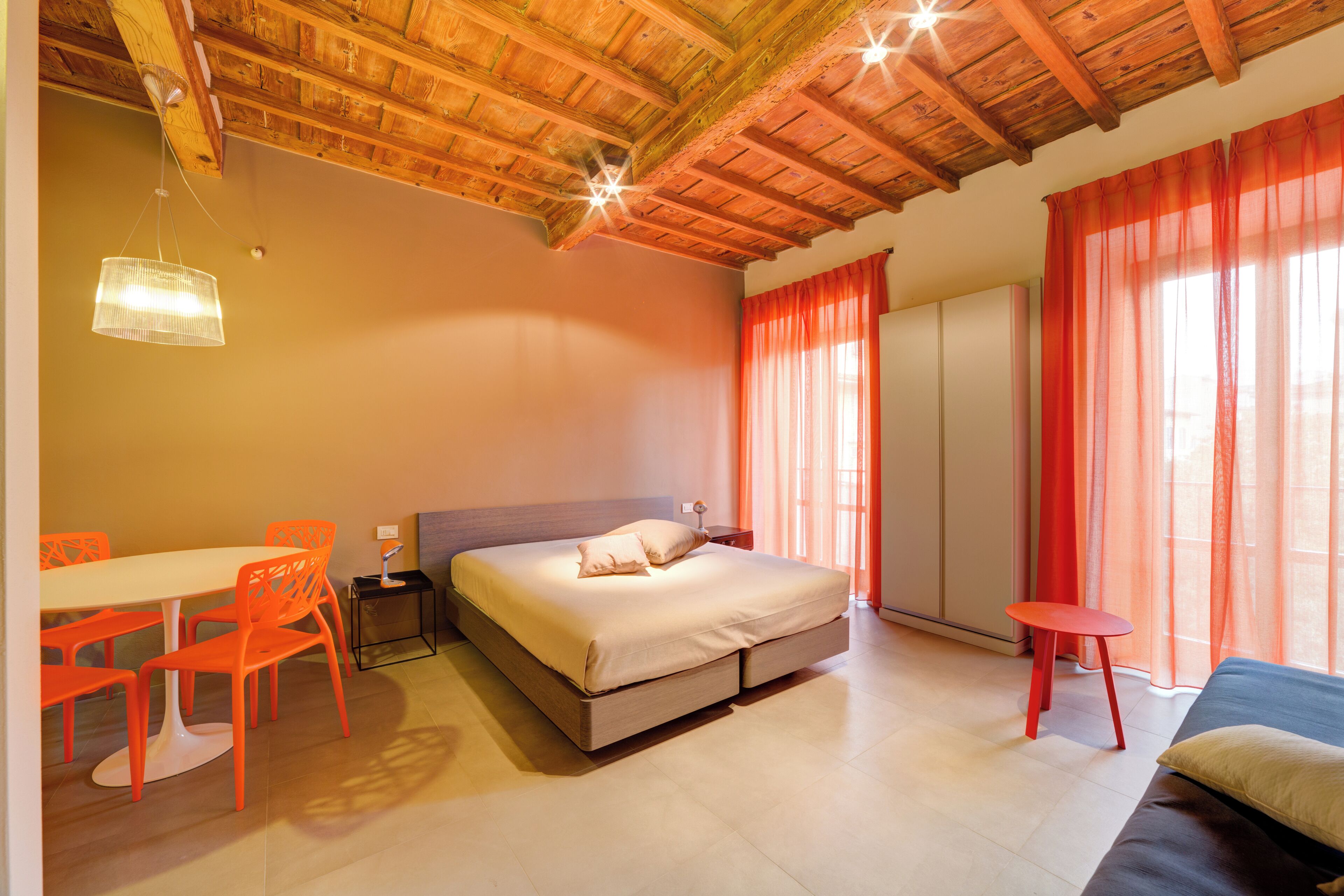 A bedroom with a wooden ceiling, a bed, a table with chairs, and a window with curtains.