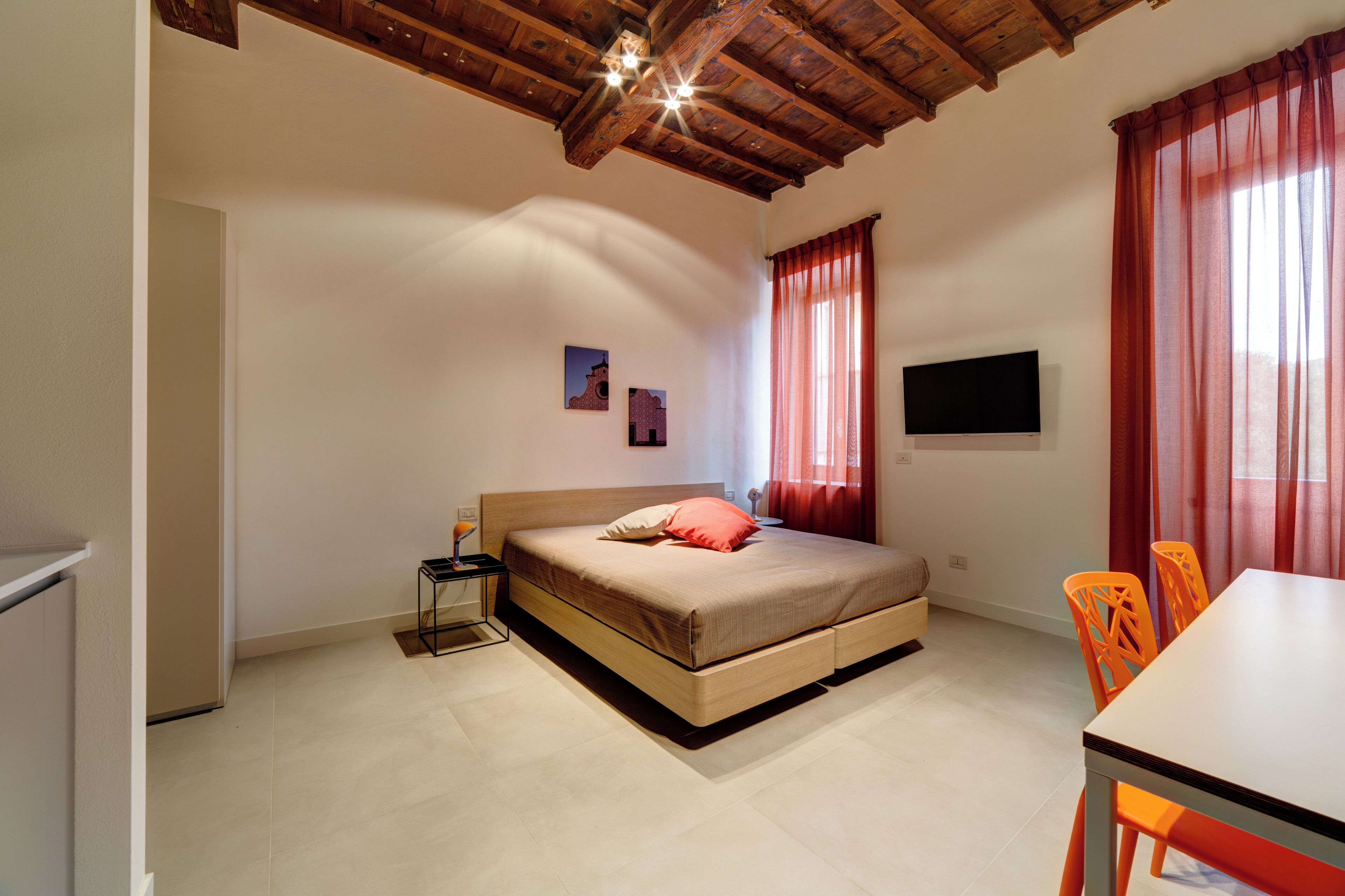 Une chambre avec un plafond en bois, un lit avec un matelas et des oreillers, une table de chevet avec une lampe, un téléviseur fixé au mur et une fenêtre avec des rideaux rouges.