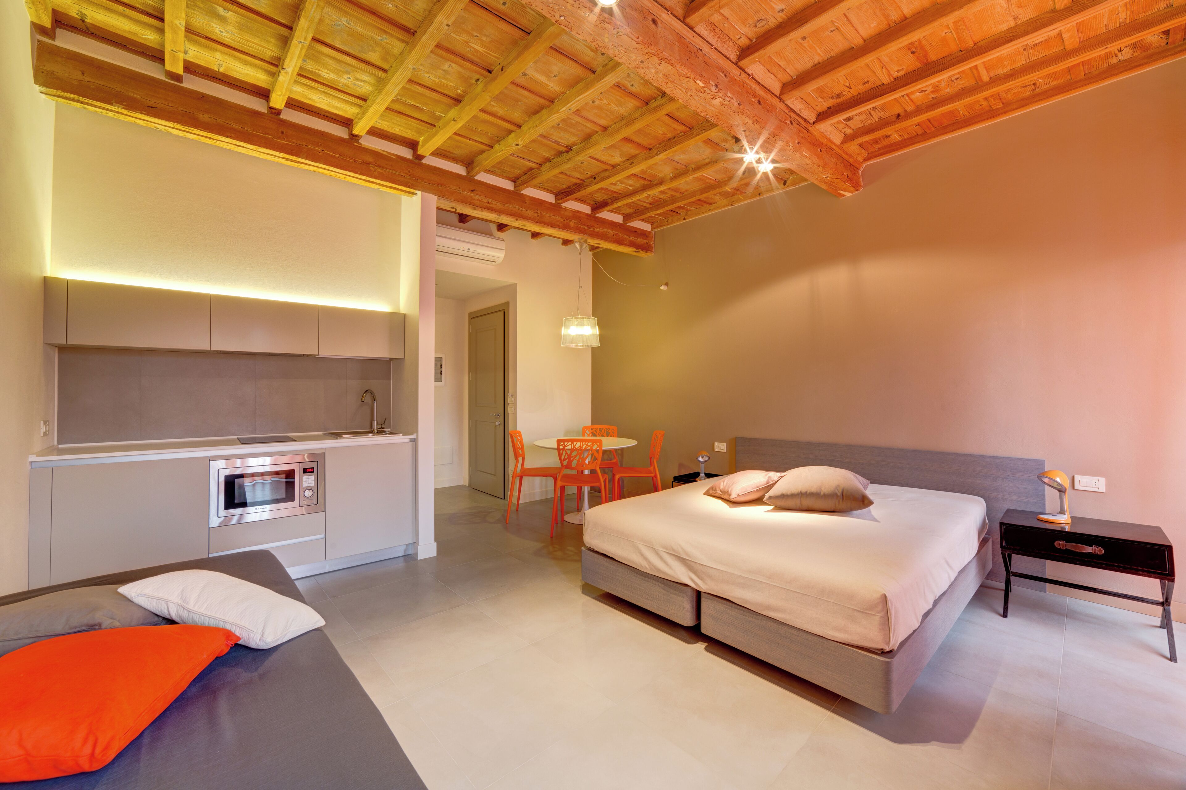 A modern bedroom with a large bed, a bedside table, a nightstand, a small kitchen area with a microwave, and a dining table with chairs.