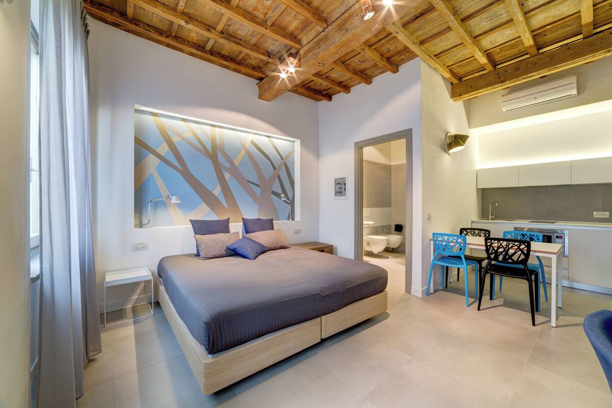 Ein modernes Schlafzimmer mit einem großen Bett, einem Schreibtisch mit Stühlen und einem durchgehenden Türspalt sichtbaren Badezimmer.