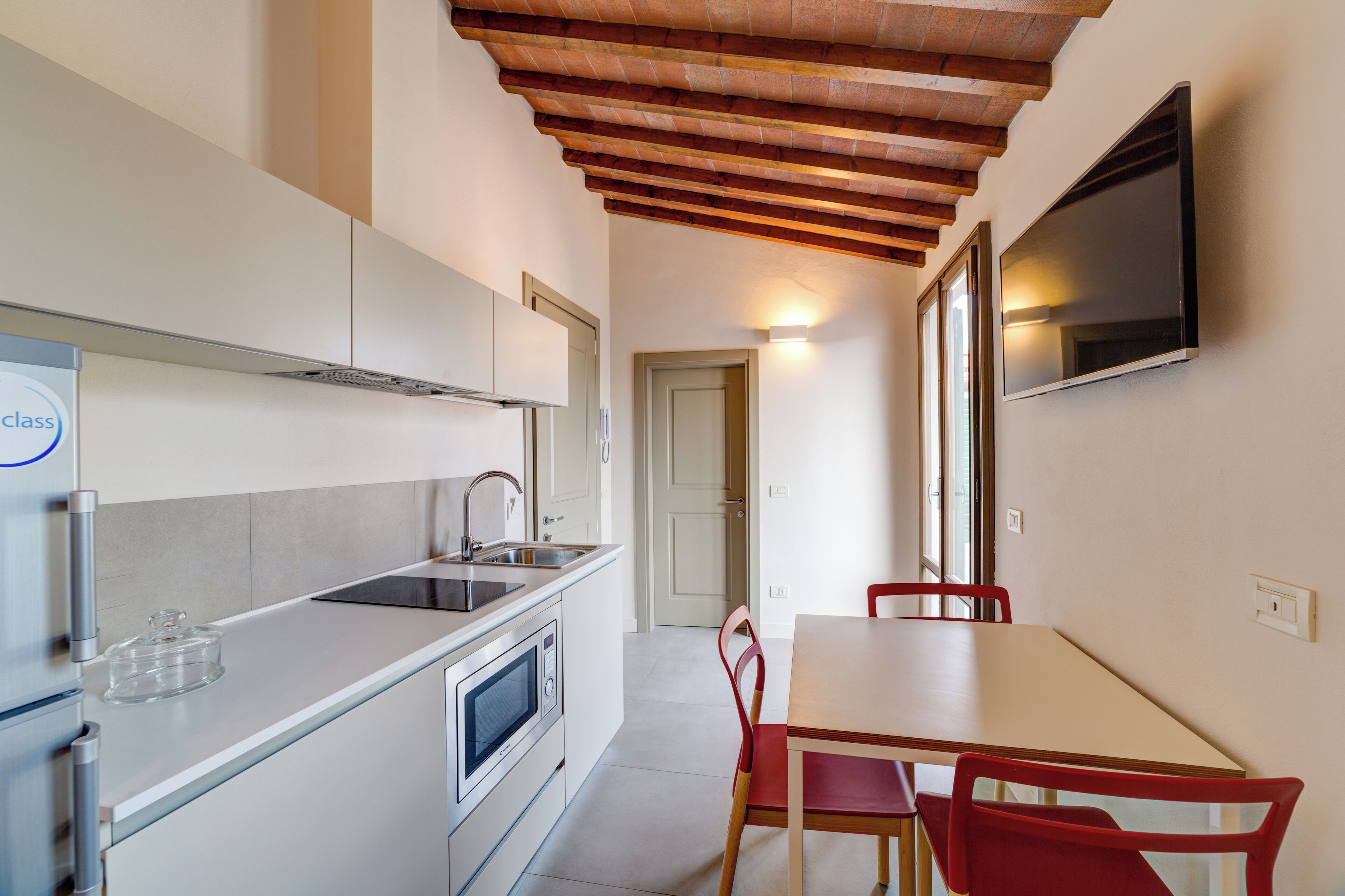 Una cocina moderna con horno empotrado, microondas y lavaplatos. Hay una mesa de comedor con sillas rojas y un televisor montado en la pared.
