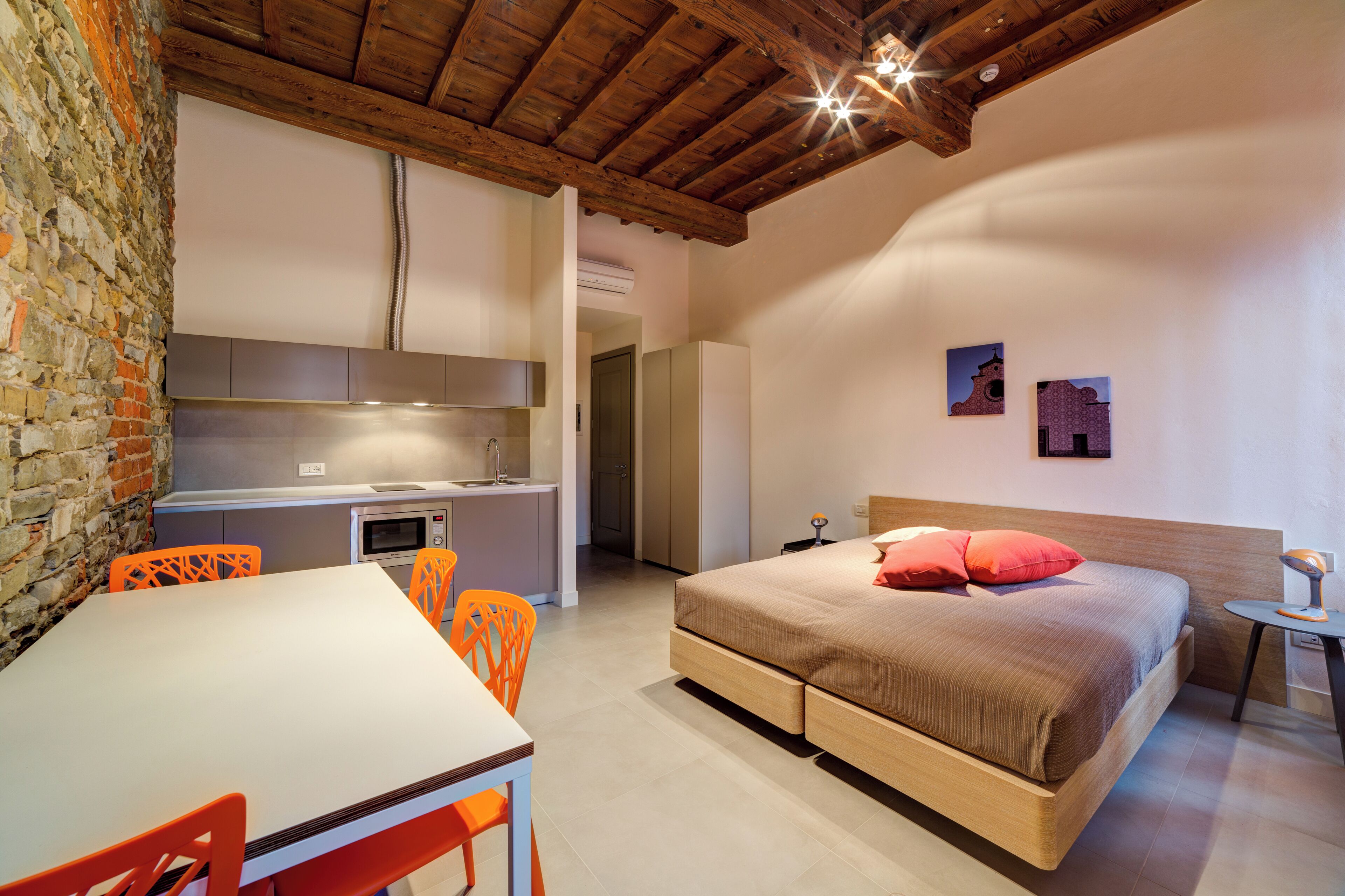 A bedroom with a bed, a desk, and a chair. There is a kitchen area with a sink and a microwave.