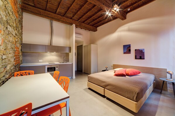 A bedroom with a bed, a desk, and a chair. There is a kitchen area with a sink and a microwave.