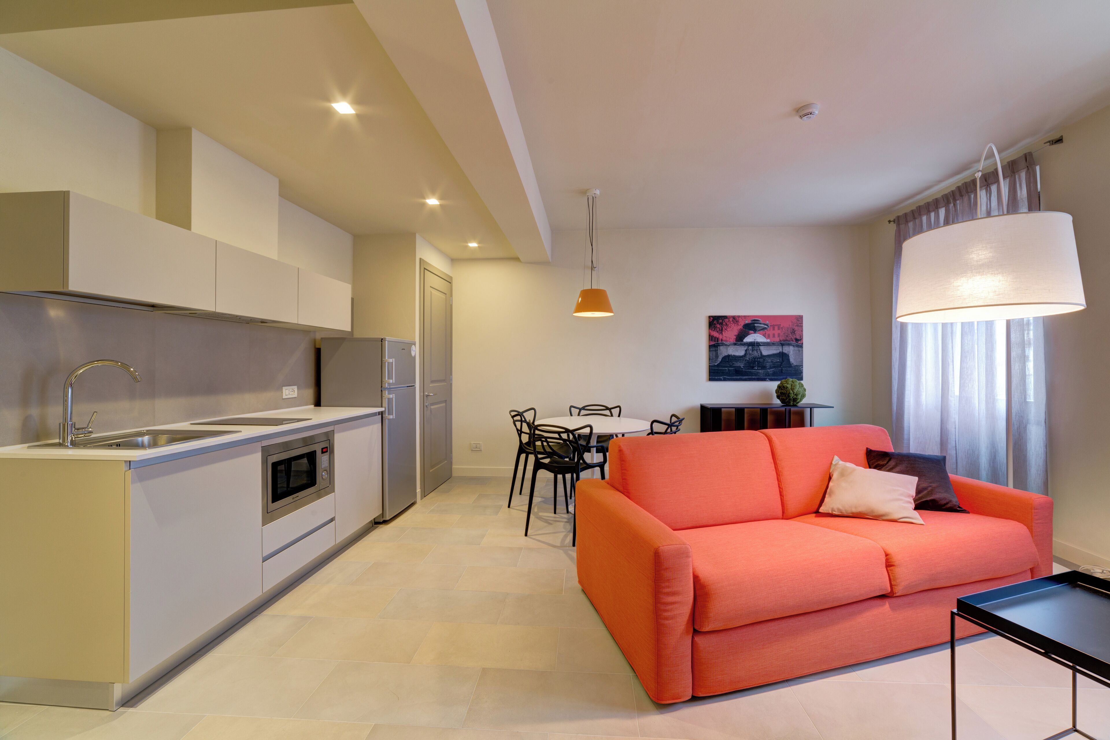A modern kitchen with a built-in oven and sink, a dining area with a table and chairs, and a bright living space with a sofa and a lamp.