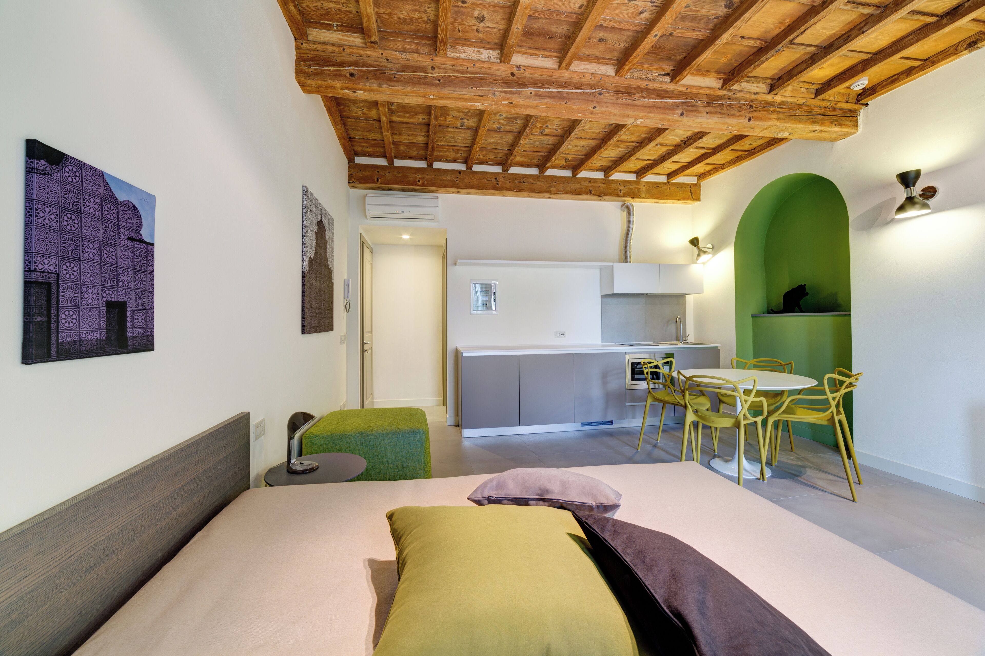 Camera da letto con soffitto in legno, un letto con molti cuscini, un poggiapiedi verde, un tavolino e una piccola cucina sullo sfondo.