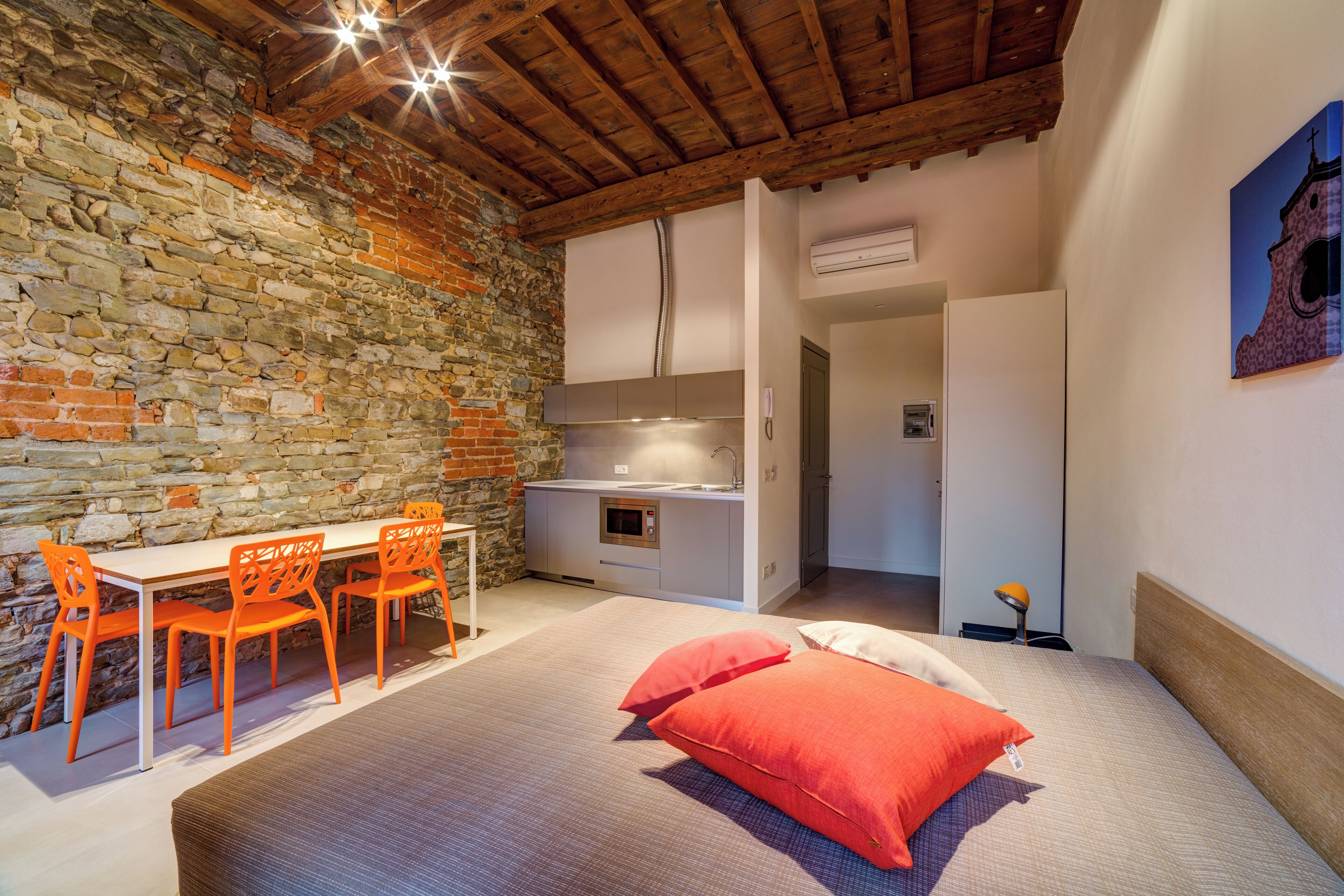 A room with a stone wall, wooden ceiling, a bed with red pillows, a dining table with orange chairs, and a kitchen area.