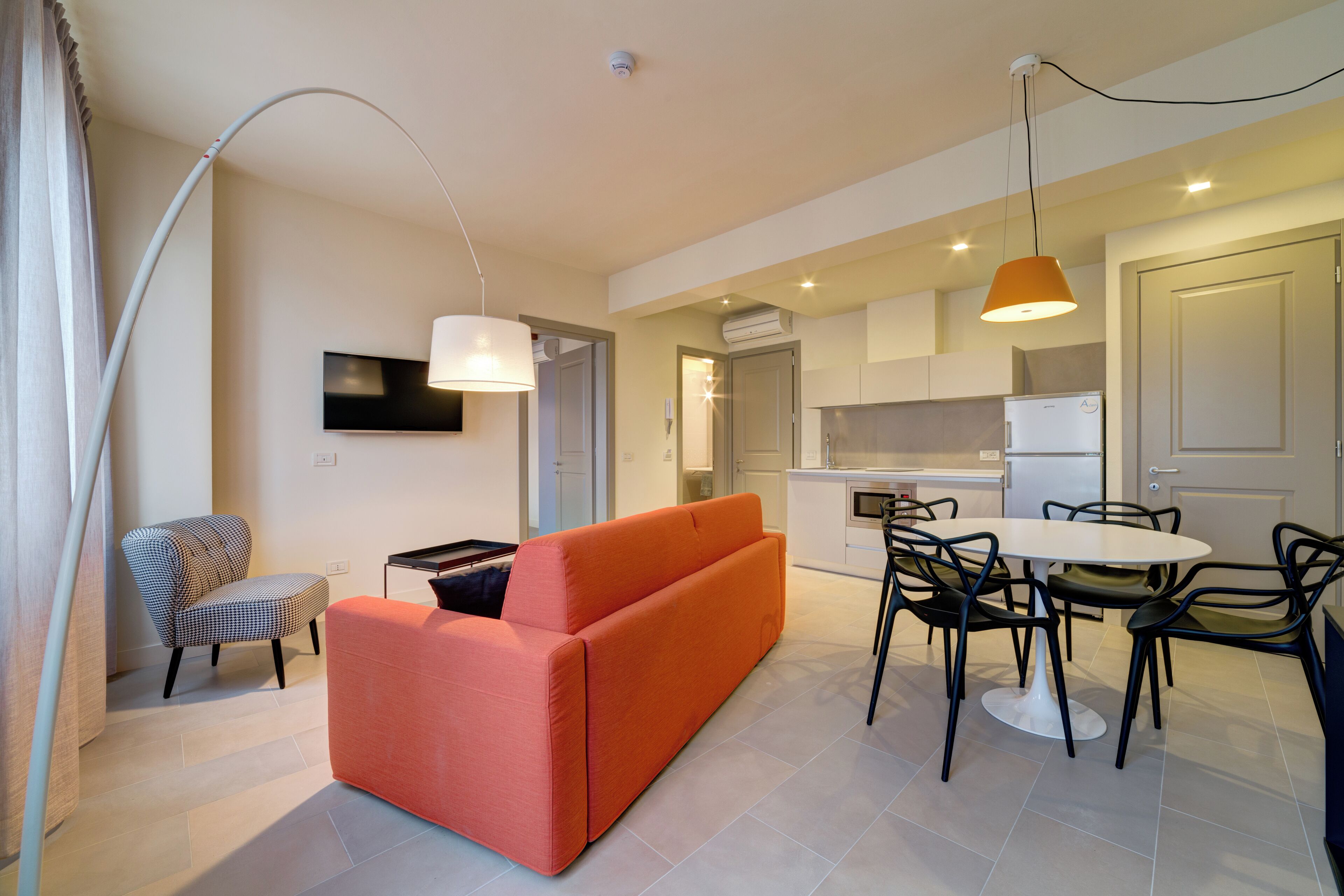 A modern living room with a sofa, a dining table with chairs, a TV mounted on the wall, and an air conditioning unit in the background.