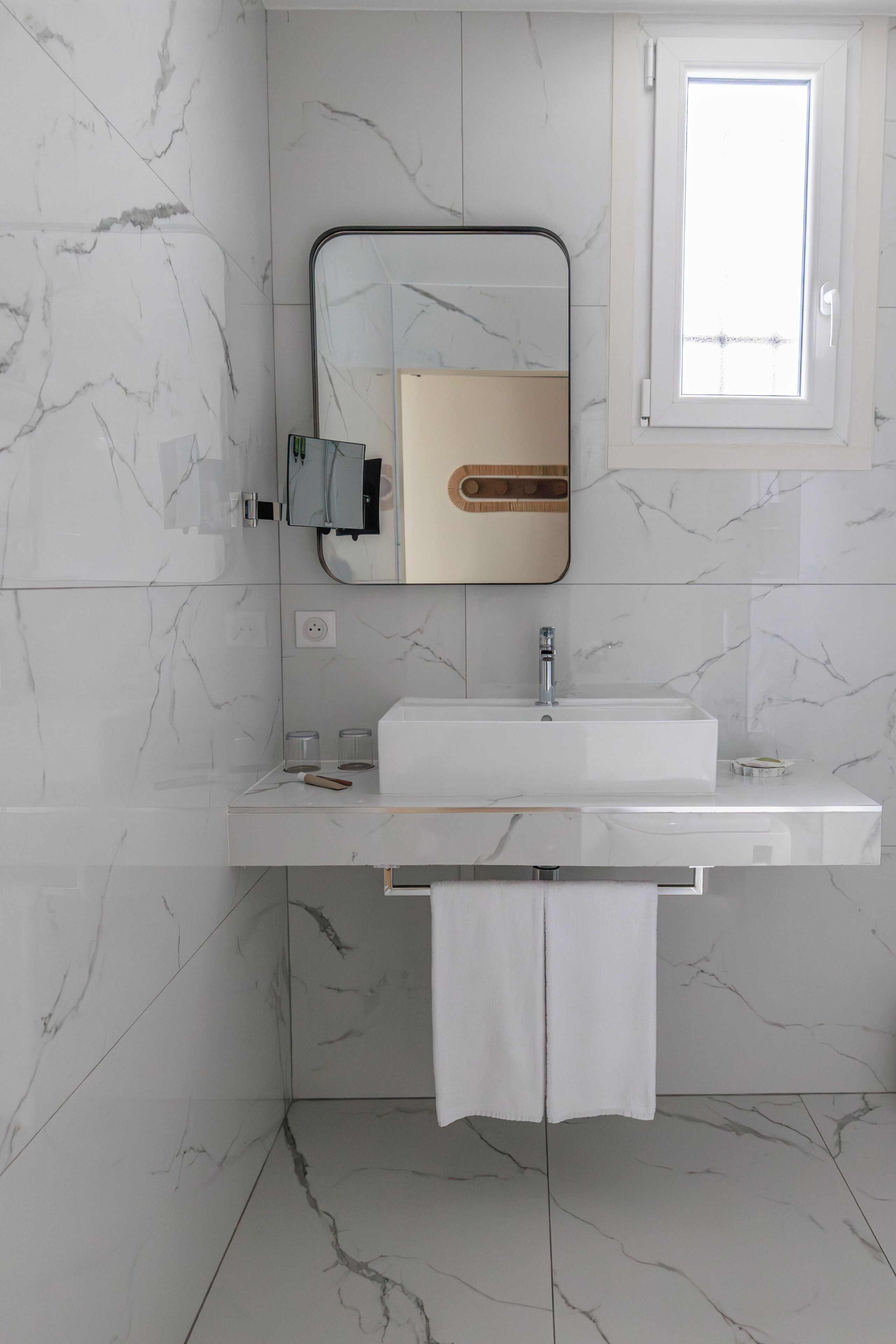 A modern bathroom with a marble-tiled wall, a white sink, a mirror, and a towel hanging on a rack.