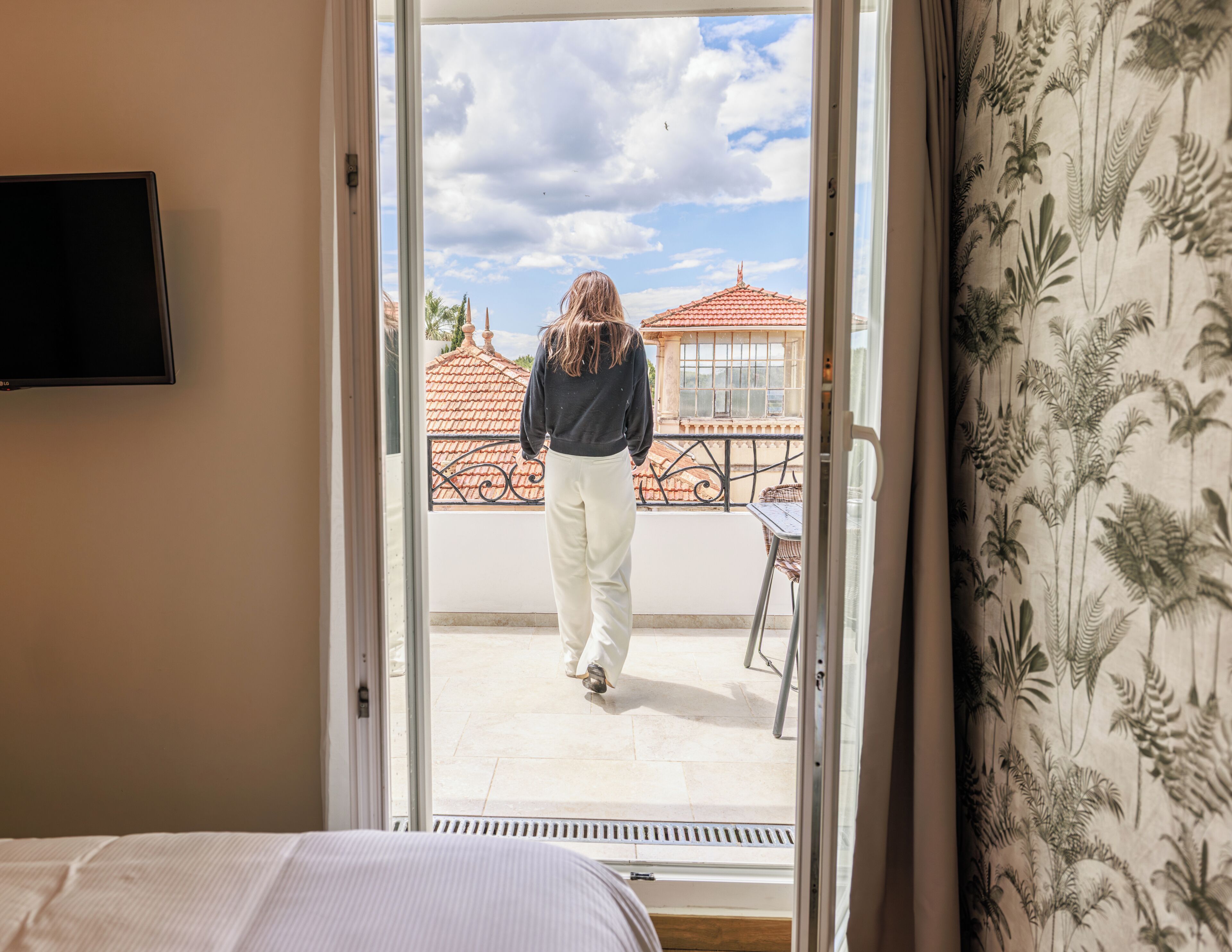A room with a bed, a television on the wall, and a view of a balcony with a person walking away.