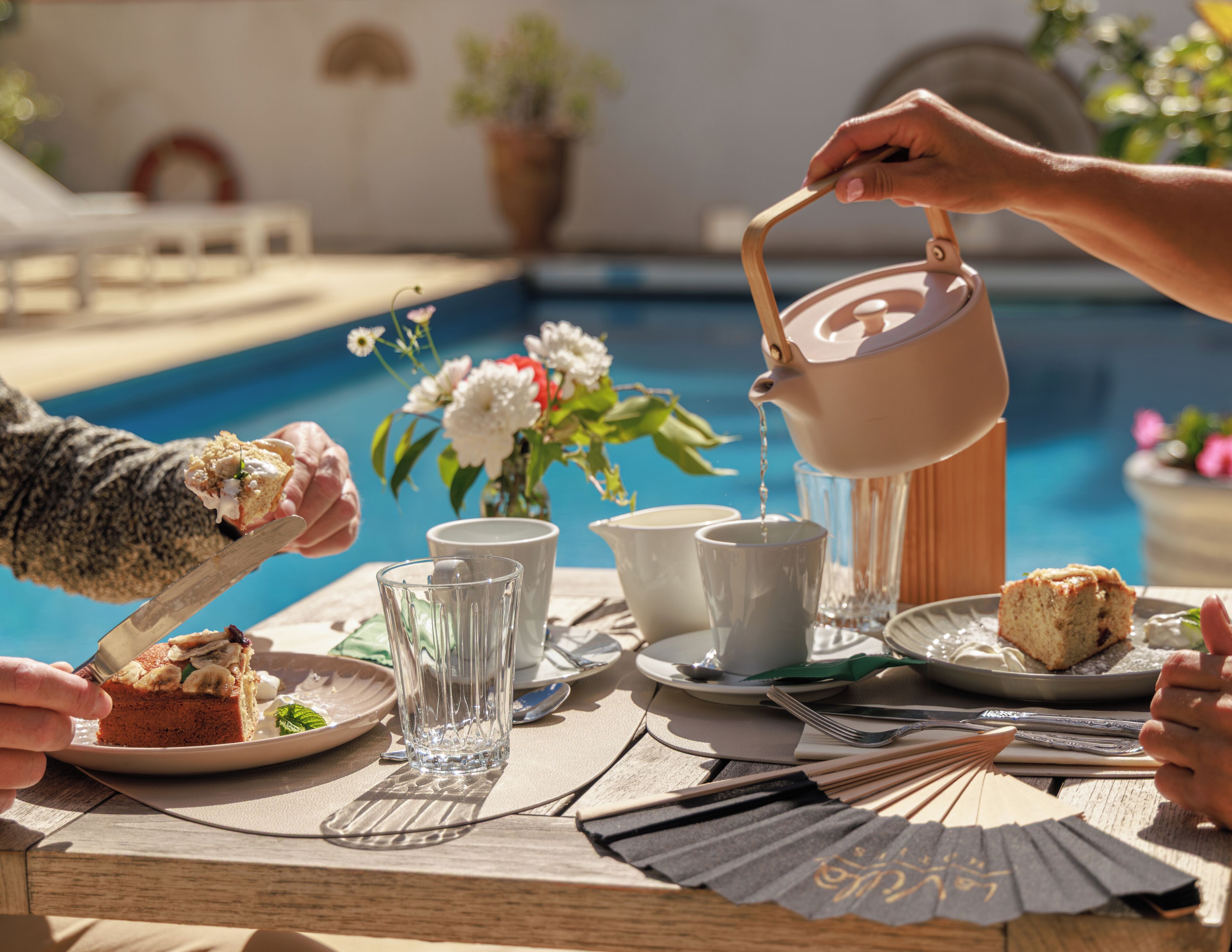 Ein Esstisch mit einer Teekanne, die Tee einschenkt, einem Teller mit Kuchen und einem Glas Wasser.