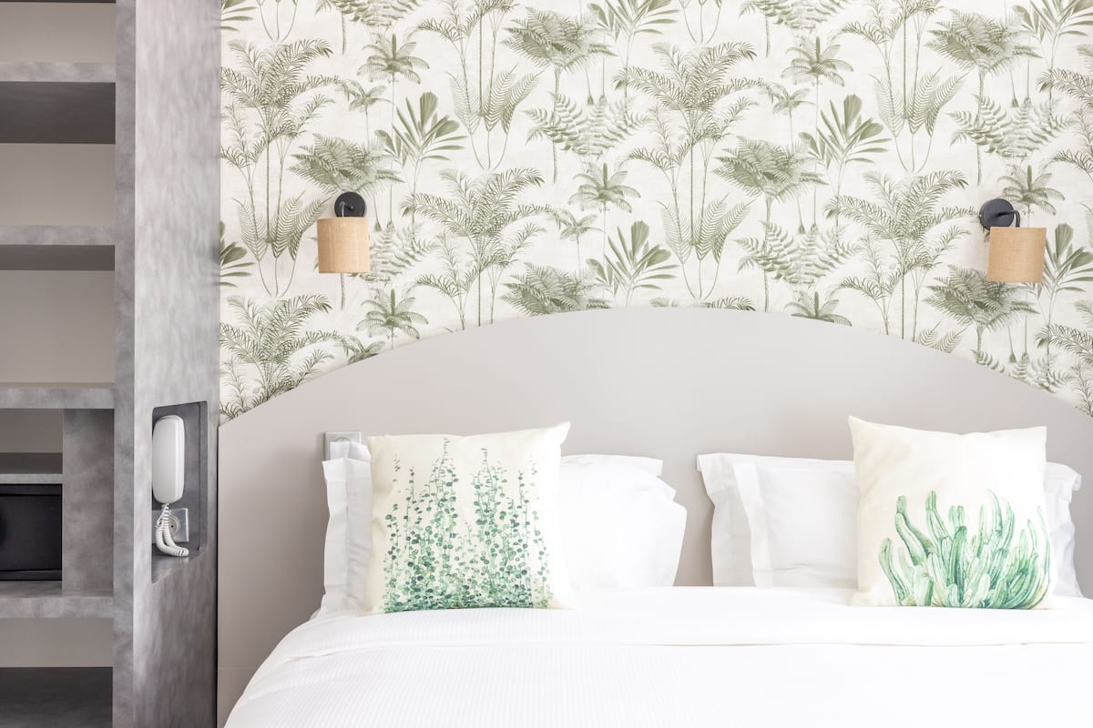 A hotel room with a bed, white bedding, a wall with a leaf pattern, and two wall-mounted lamps.