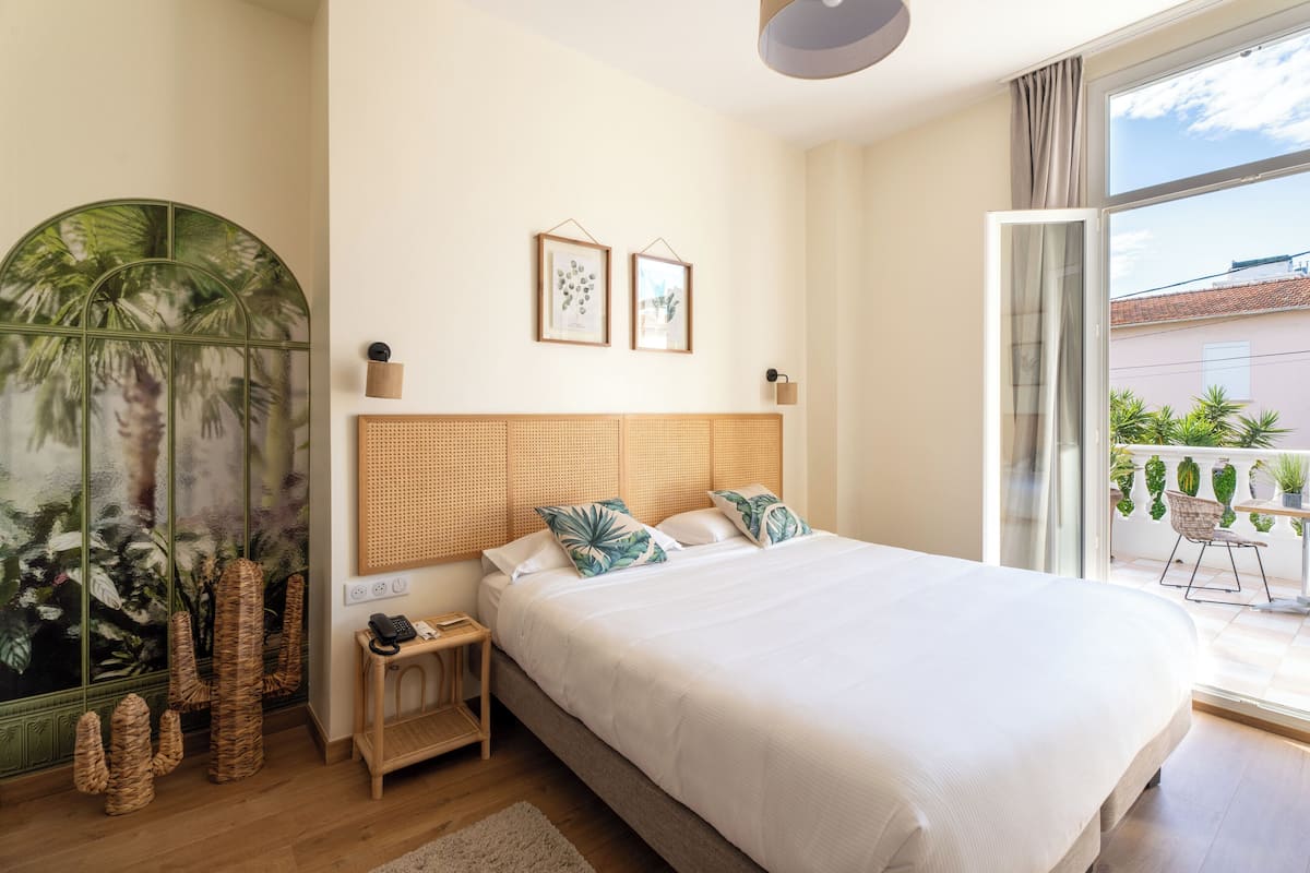 A bedroom with a large bed, a nightstand, a wicker chair, a wall-mounted lamp, and a window with a view of the outside.