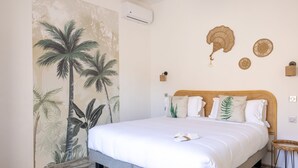 A bedroom with a large bed, a wall mural of palm trees, a ceiling fan, and a small wicker side table. - Hotel La Villa Juan Beach (Antibes)