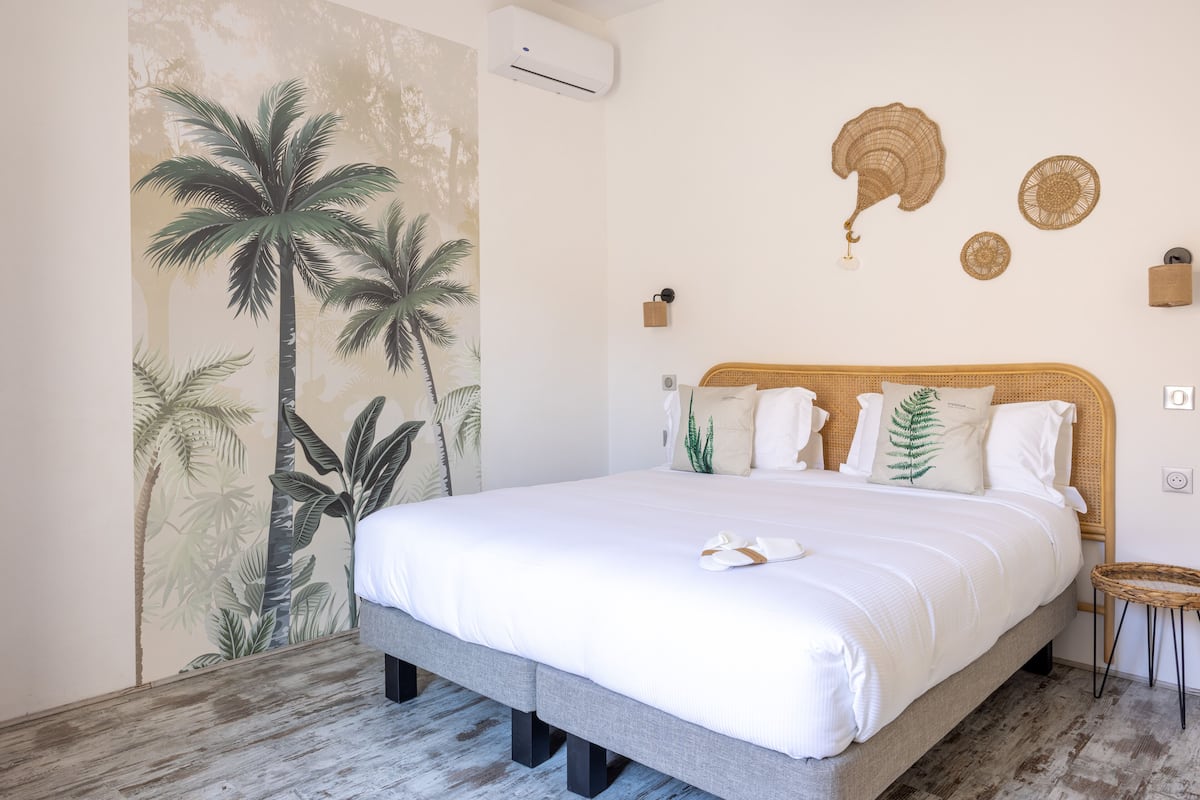 A bedroom with a large bed, a wall mural of palm trees, a ceiling fan, and a small wicker side table.