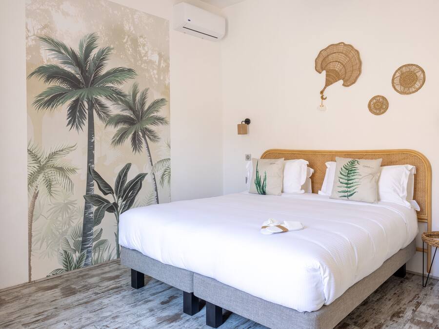 Un dormitorio con una cama grande, un mural en la pared con palmeras, un ventilador de techo y una pequeña mesita de mimbre.