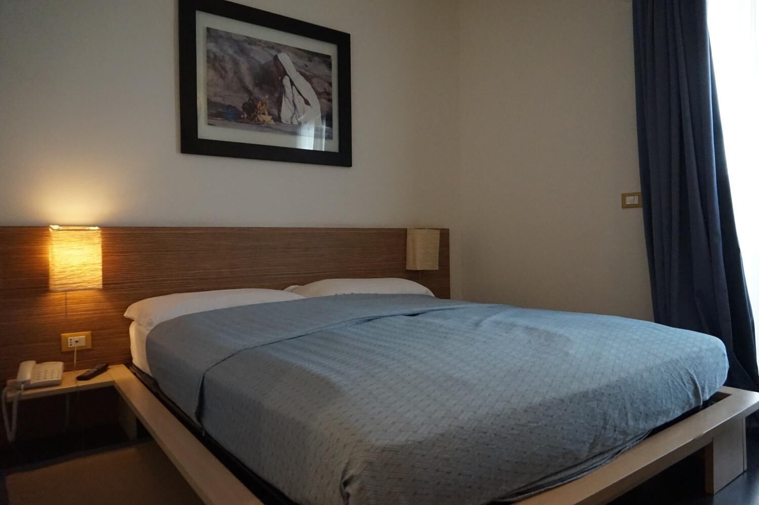 Una cama bien hecha con una manta azul, cabecera de madera, lámpara de noche y un cuadro en la pared.