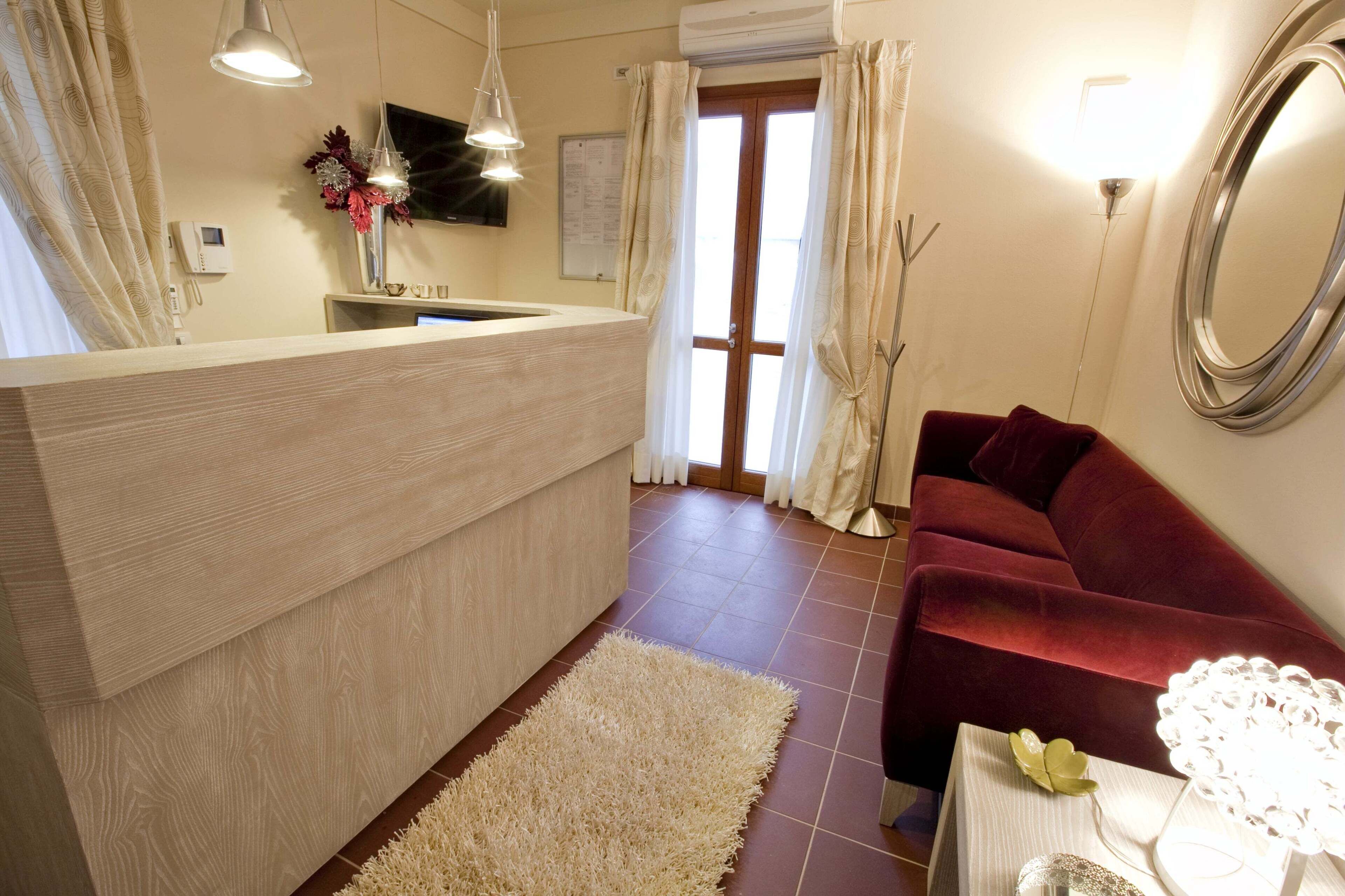 A modern hotel room with a wooden reception desk, a red sofa, a white rug, and a large mirror on the wall.