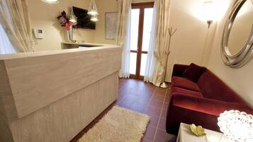 A modern hotel room with a wooden reception desk, a red sofa, a white rug, and a large mirror on the wall.