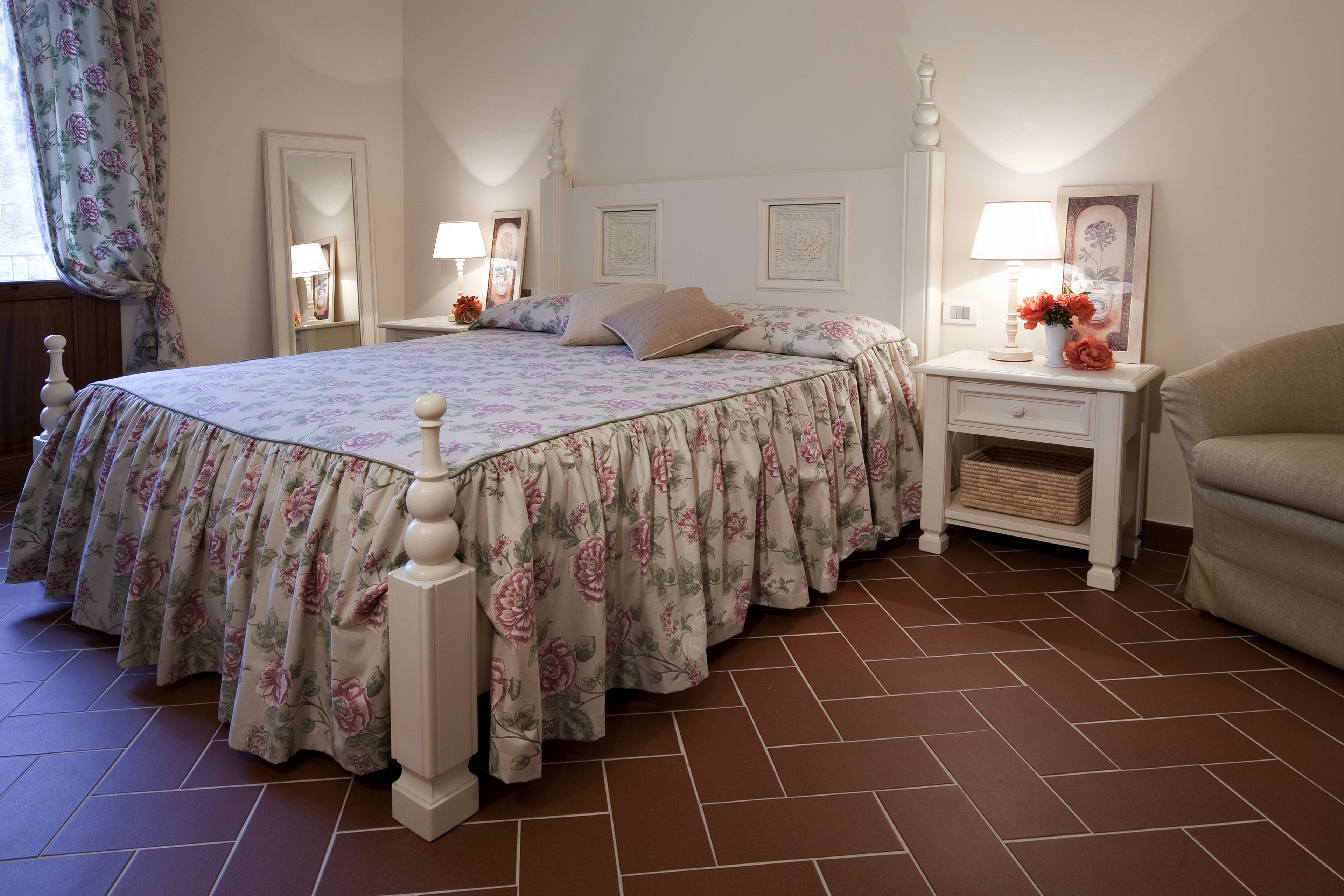 Um quarto com edredom floral, cabeceira branca, mesa de cabeceira com abajur e uma cadeira.