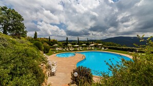 A large outdoor swimming pool surrounded by lounge chairs and umbrellas, with a scenic view of hills and a cloudy sky. - Tenuta Quadrifoglio (Gambassi Terme)