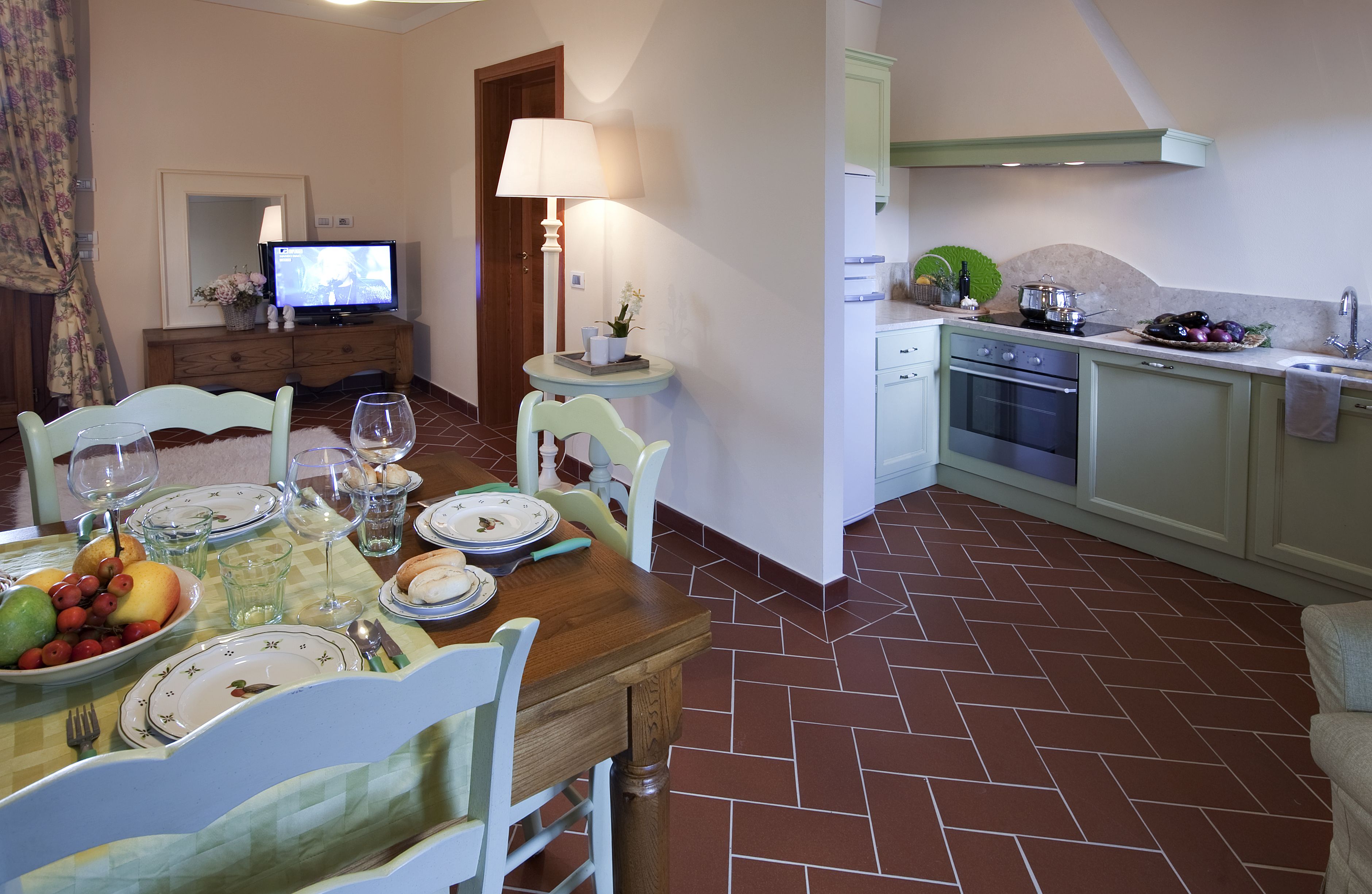 A dining area with a wooden table set for four, a television in the background, and a kitchen with a stove and sink.