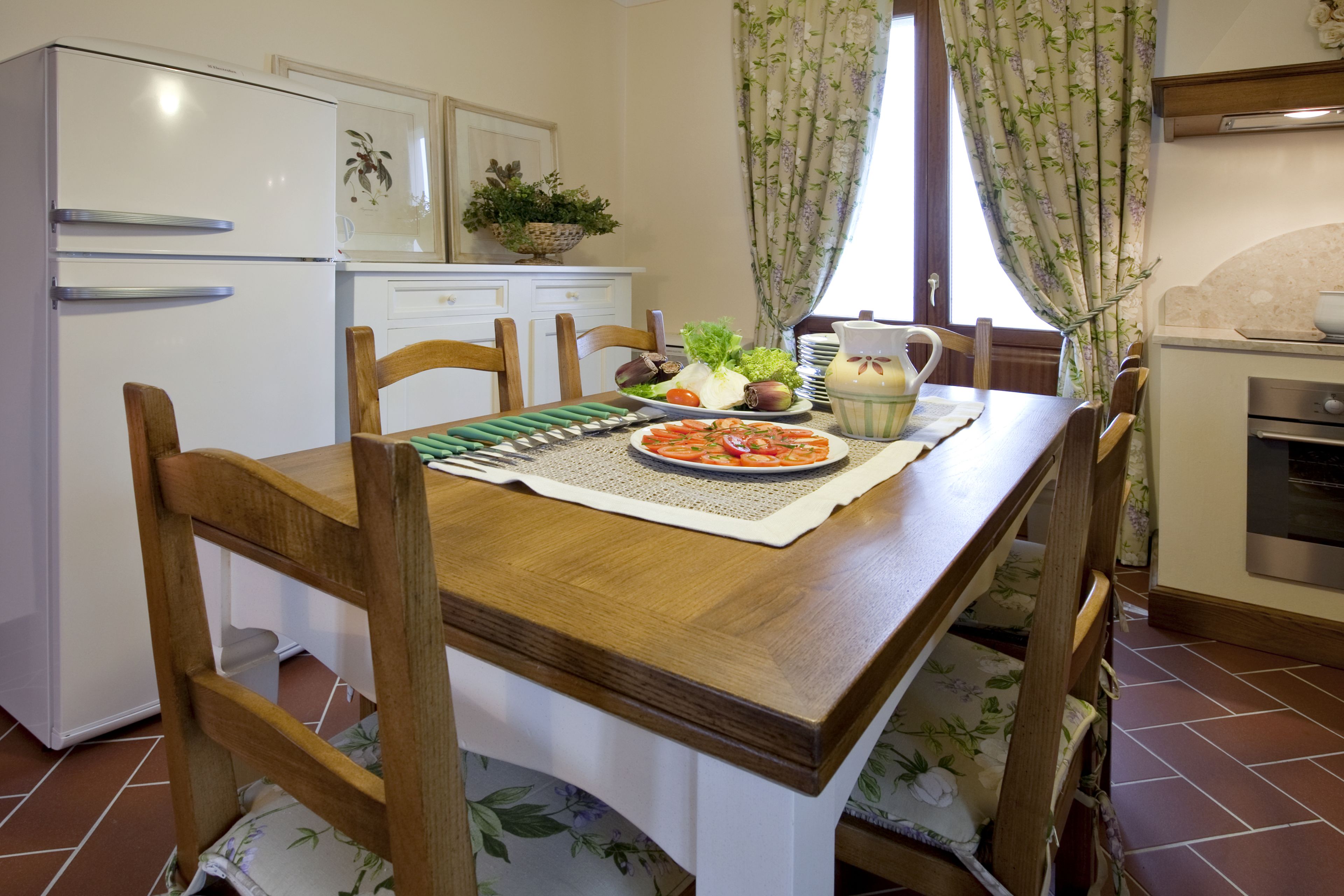 Una cocina con una mesa de madera, sillas, un refrigerador, un lavamanos y una ventana con cortinas.