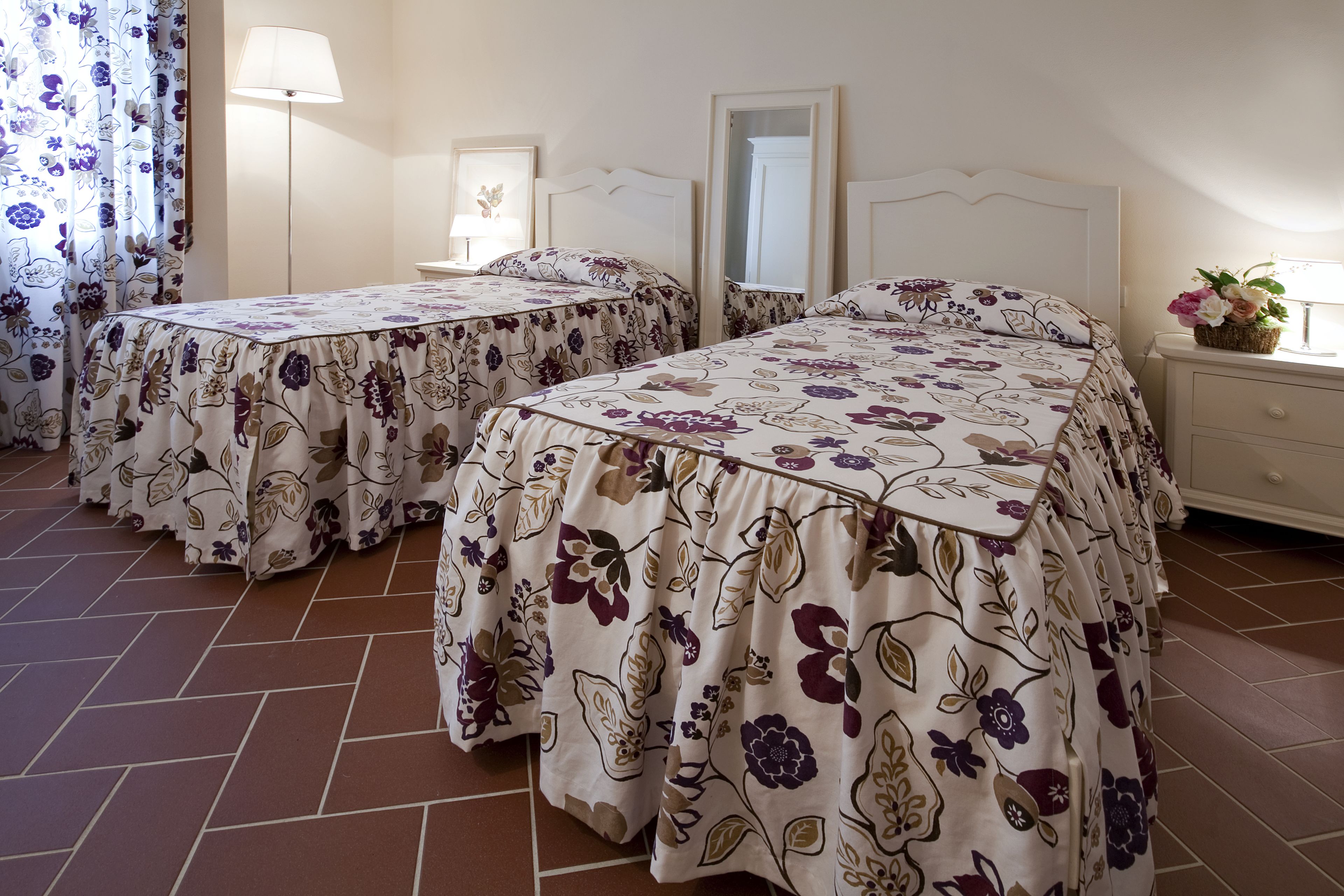 Habitación de hotel con dos camas, cada una con cubrecamas florales, una mesita de noche con lámpara, un espejo y una pequeña planta en el tocador.