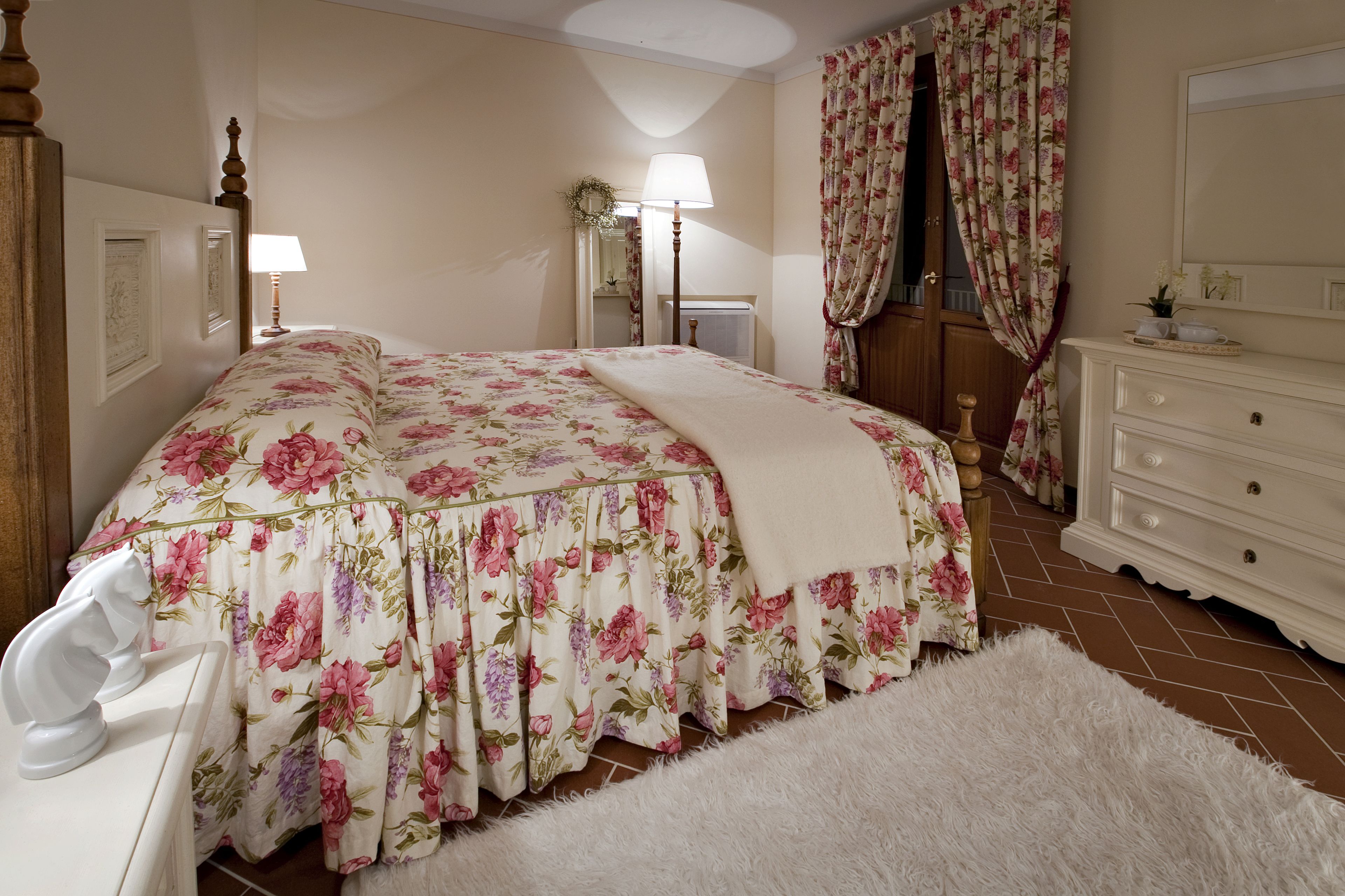 Een slaapkamer met een bloemenbedekking, een witte kapstok, een spiegel en een witte stoel.