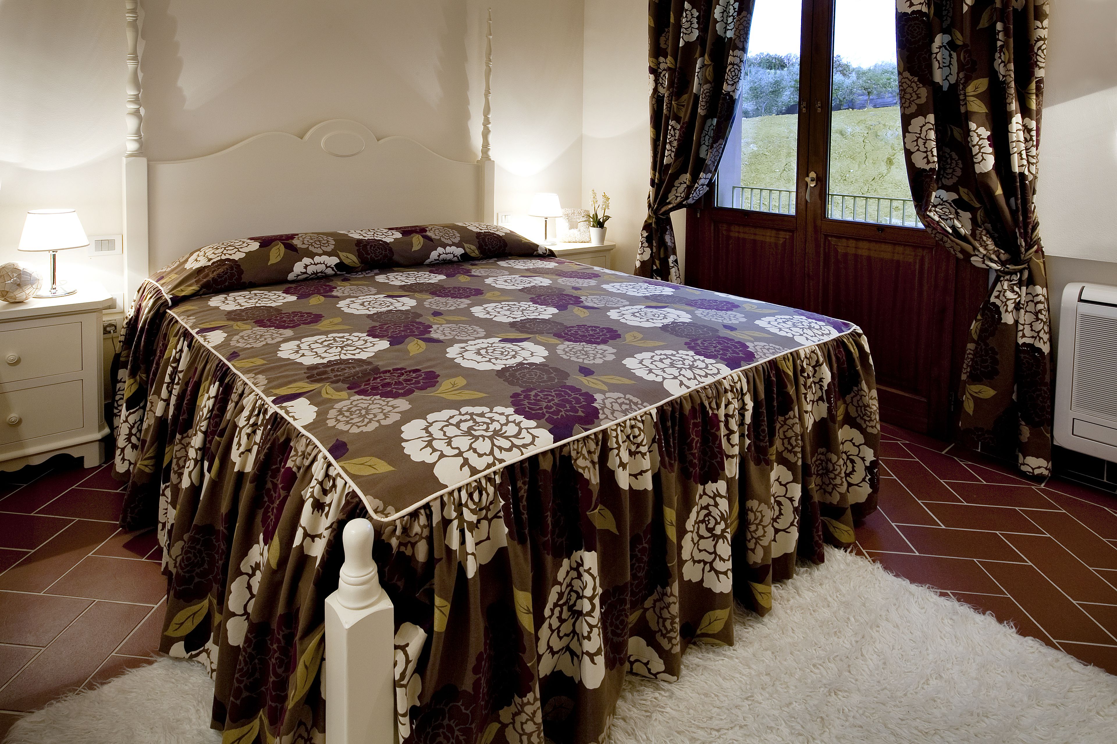 Ein Schlafzimmer mit Bett, Nachttisch, Lampe, Fenster mit Vorhängen und Heizkörper.