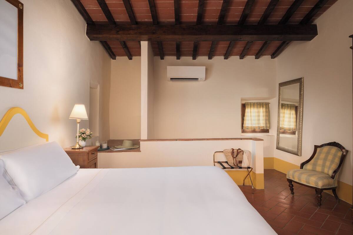 Una habitación de hotel con cama, mesita de noche, lámpara, silla, ventana con cortinas y un equipo de aire acondicionado.