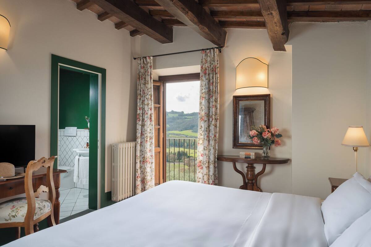 Un dormitorio con cama, escritorio, silla, radiador, ventana con cortinas, lámpara y espejo enmarco.