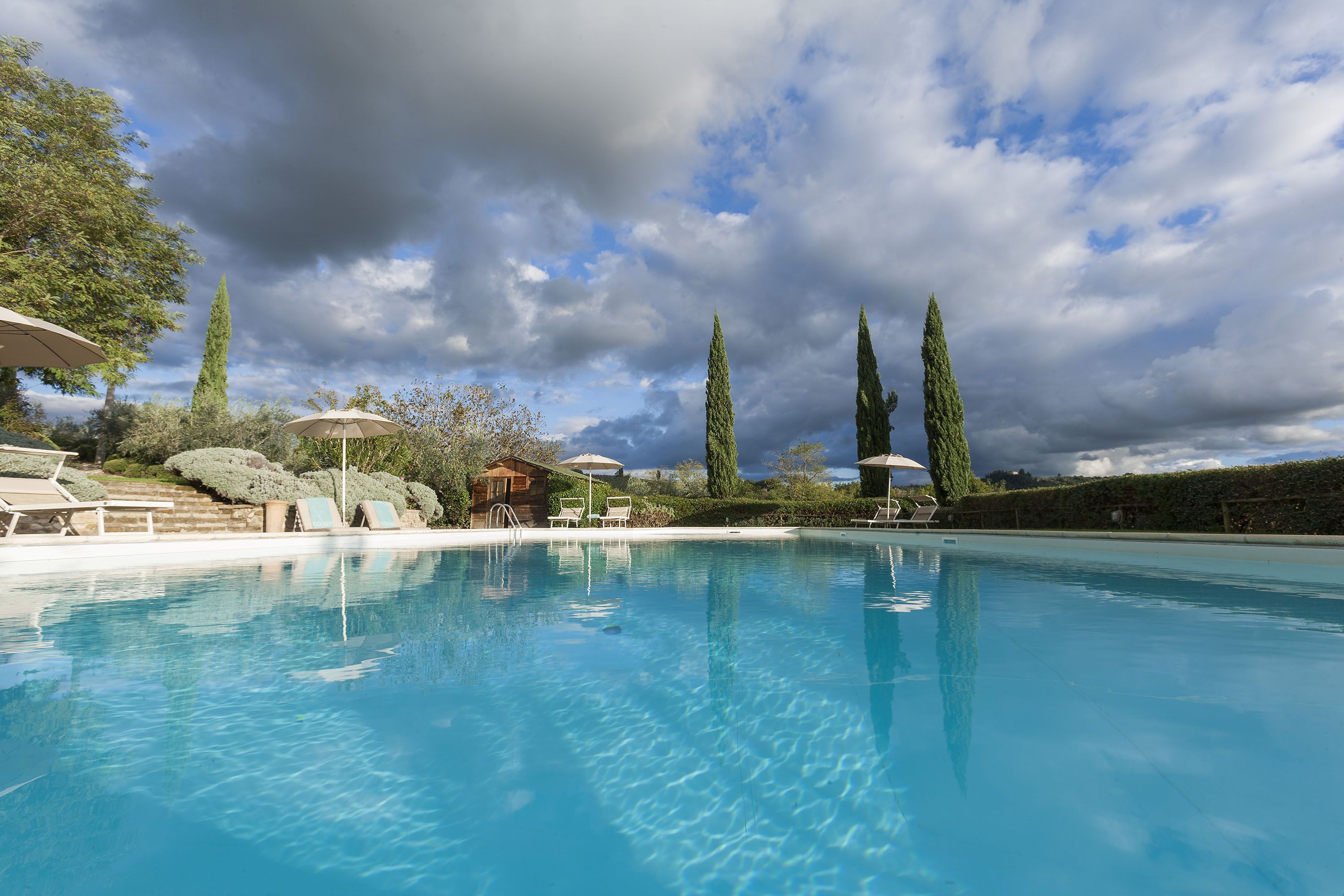 Una piscina limpida con lettini e ombrelloni, circondata da alberi alti e un cottage in legno.