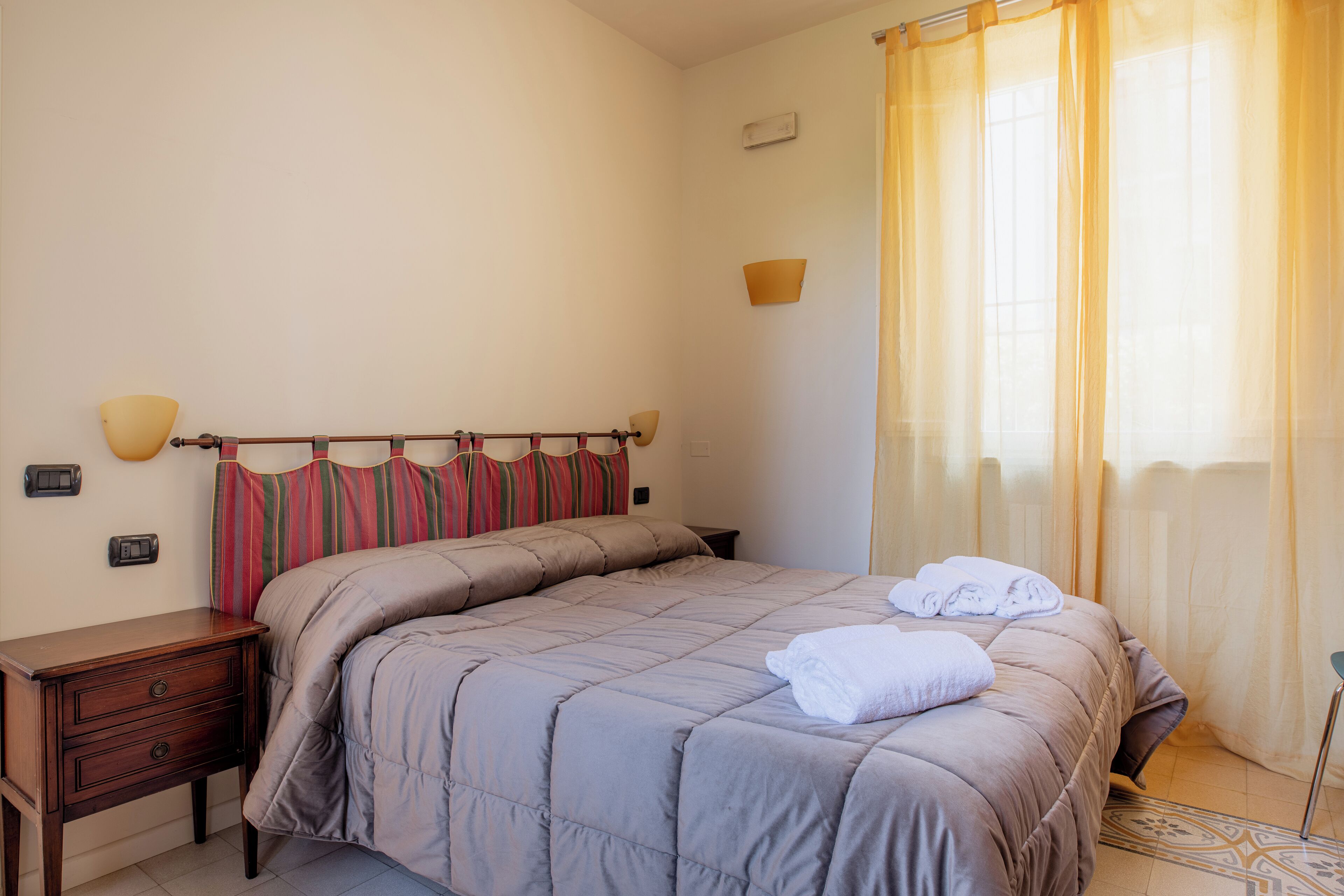 Una cama bien hecha con un cabecero a rayas, una mesita de noche con una lámpara y una ventana con cortinas translúcidas.