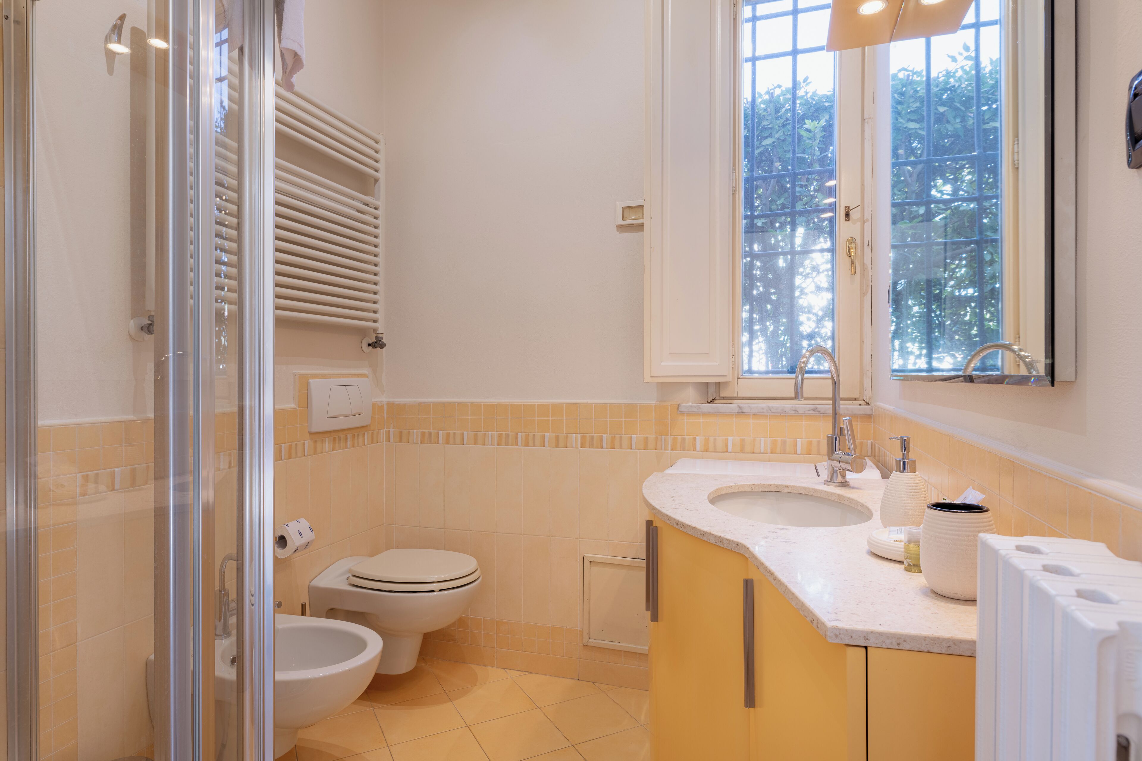 A bathroom with a glass shower enclosure, a toilet, a bidet, a sink with a mirror, and a window providing natural light.