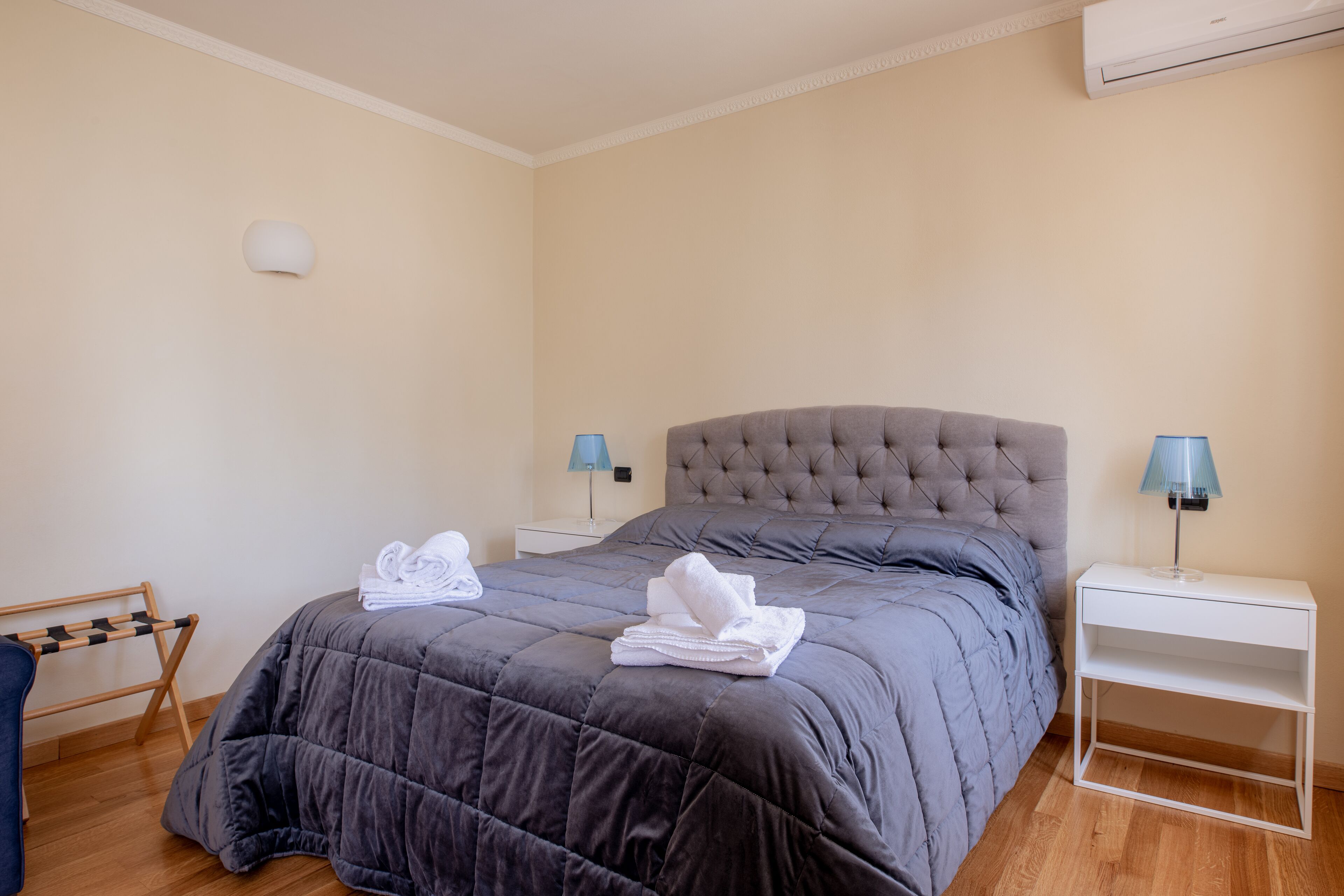 Una cama bien hecha con edredón gris, dos mesitas de noche con lámparas y una silla de madera.