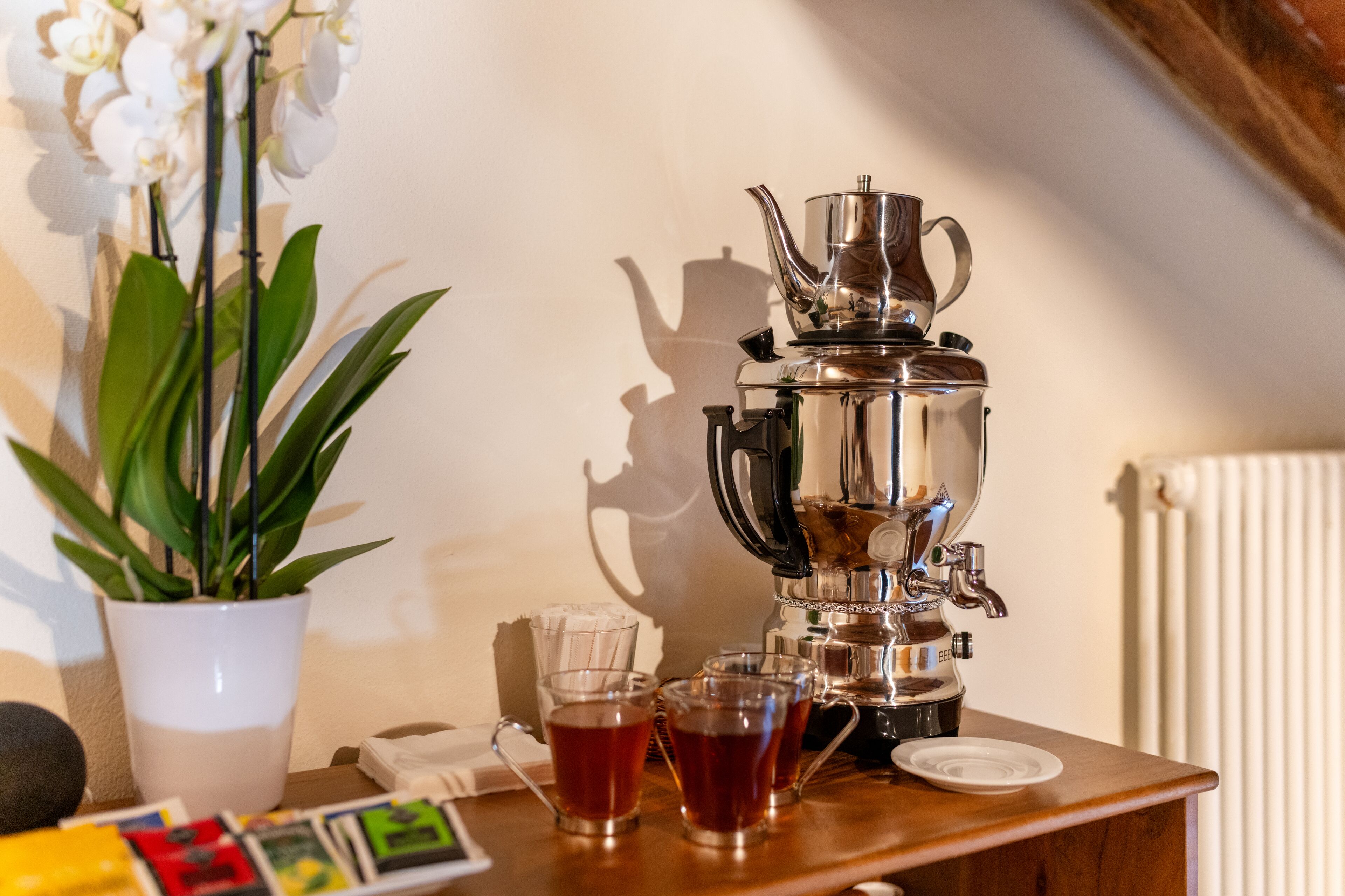 Eine Kaffeemaschine auf einem Holztisch mit verschiedenen Teegläsern, einer Topfpflanze und einem Heizkörper im Hintergrund.
