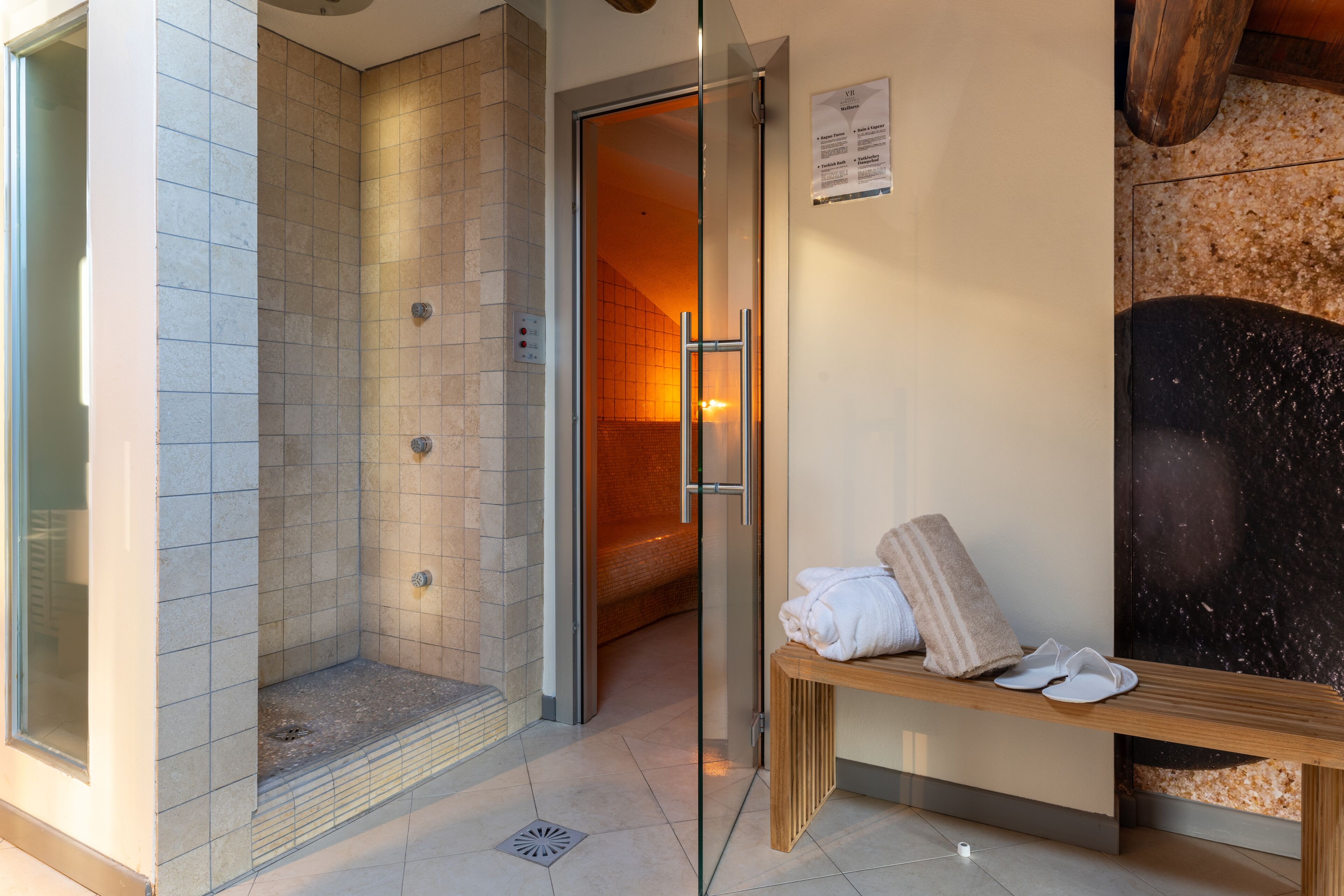 A modern bathroom with a glass-enclosed shower, a bench with towels, and a tiled floor.