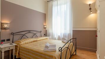 Un letto rifatto con cura, lenzuola a righe, un comodino con una lampada e una finestra con tende leggere.