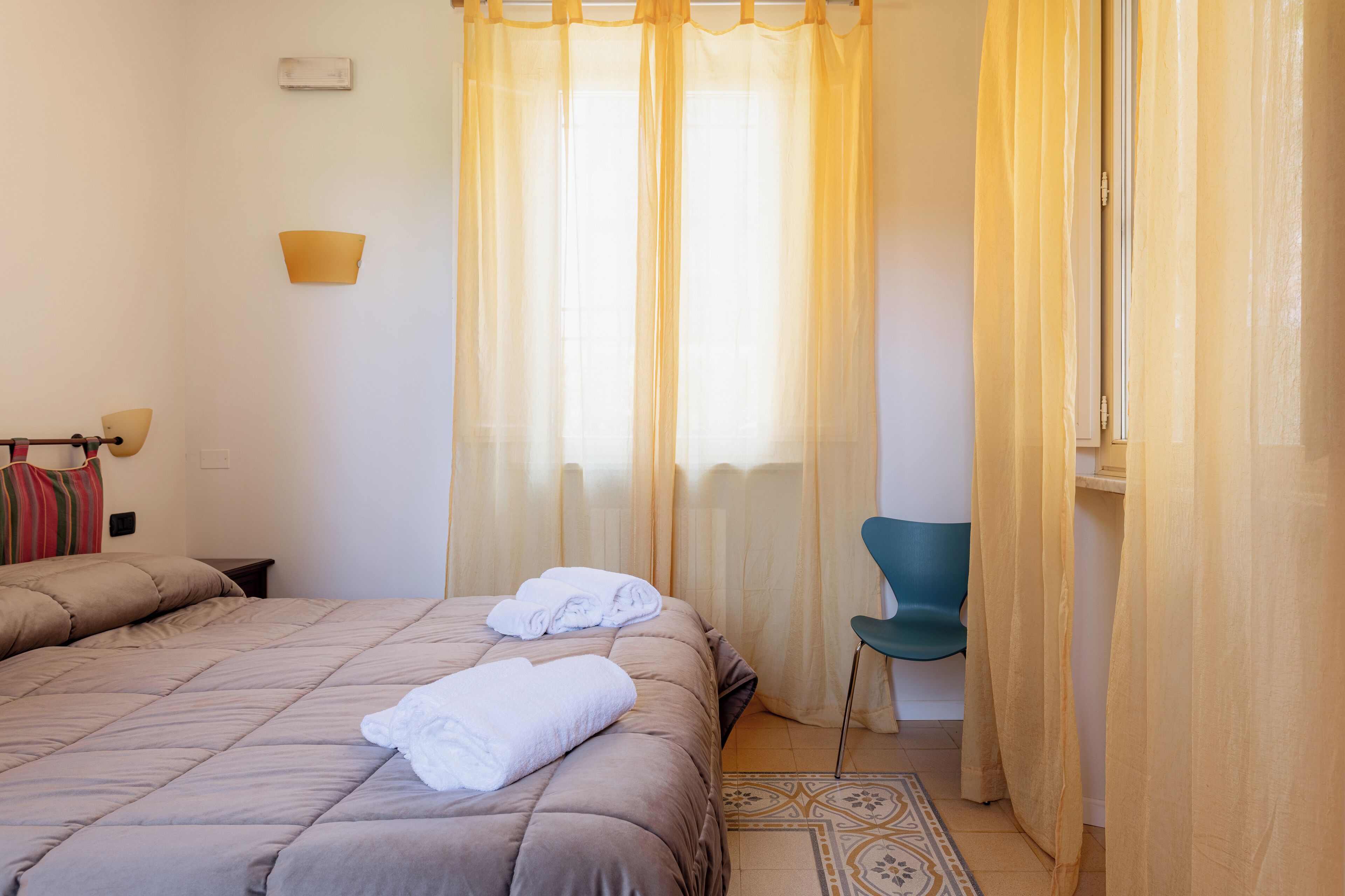 Una camera d'albergo con due letti, una sedia e una finestra con tende leggere.
