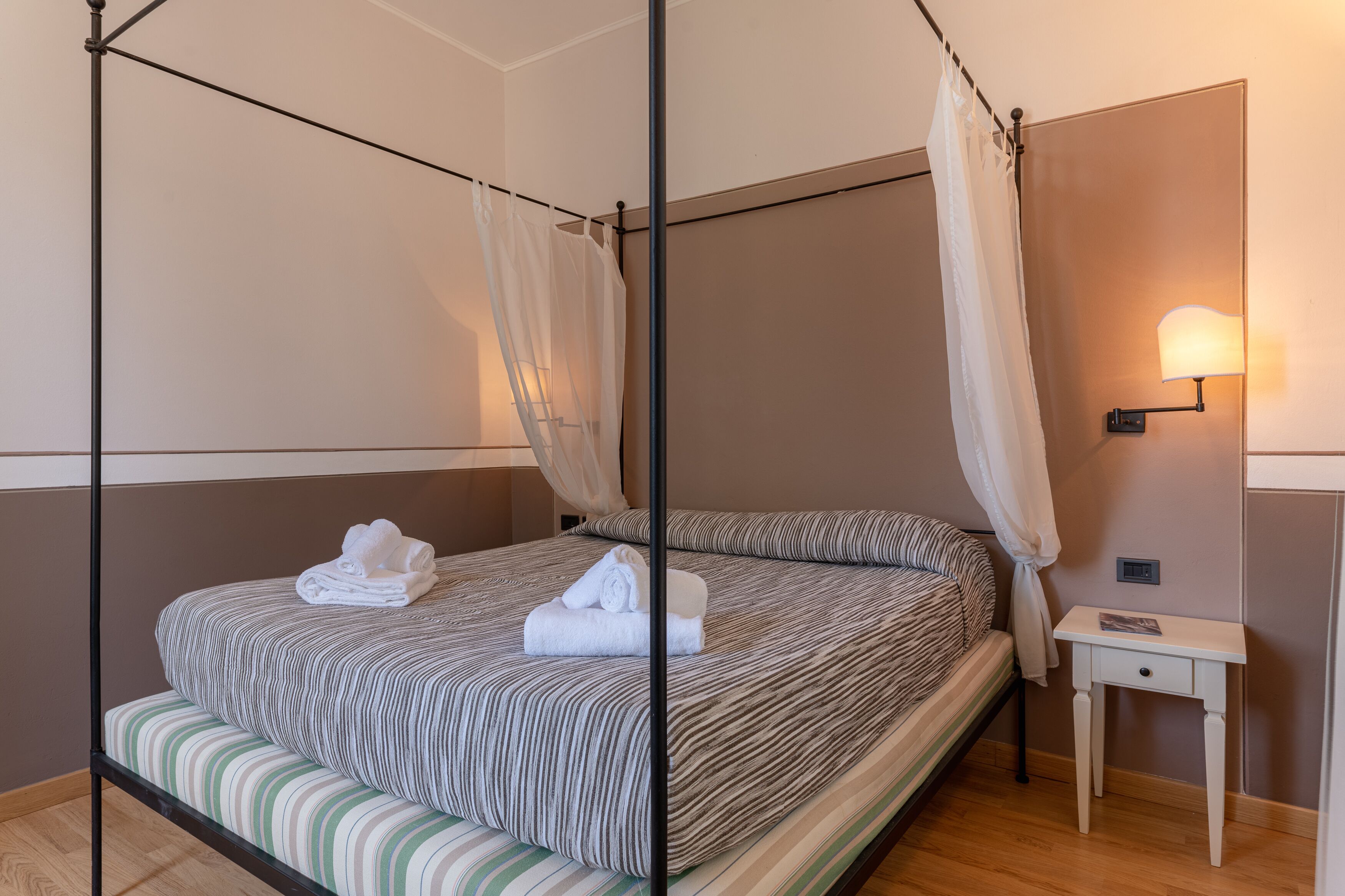 A four-poster bed with a striped bedspread and white towels, a bedside table with a lamp, and a wooden floor.