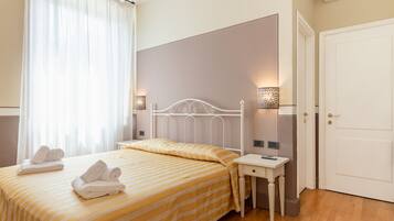 A hotel room with two beds, a bedside table, a nightstand, a door, and a window with curtains.