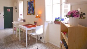 Une pièce avec une table et des chaises blanches, un tableau au mur, une lampe et une fenêtre ornée de fleurs.