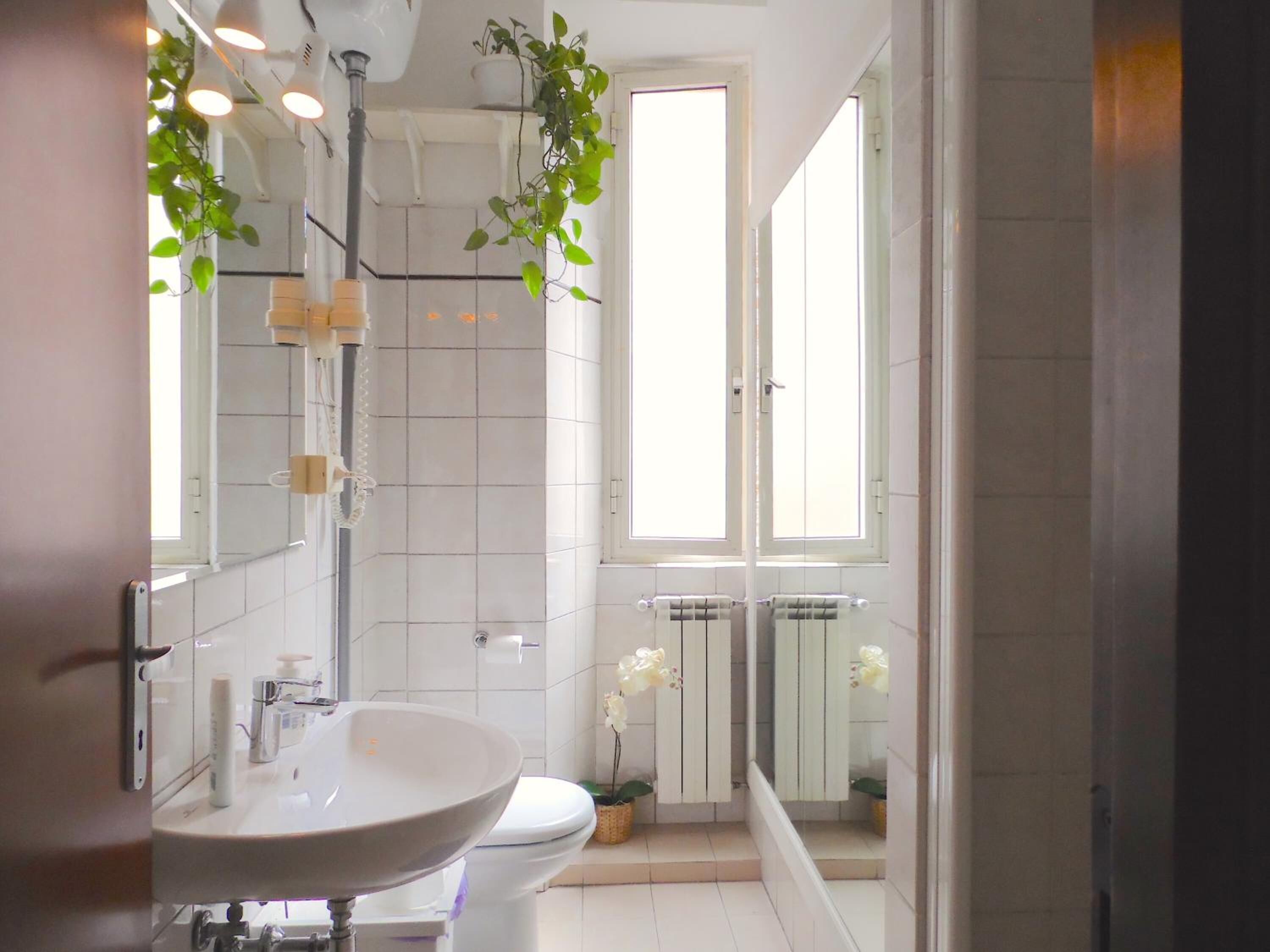 Un baño moderno con lavamanos, inodoro y ducha blancos. Hay plantas en macetas y un ventanal que proporciona luz natural.