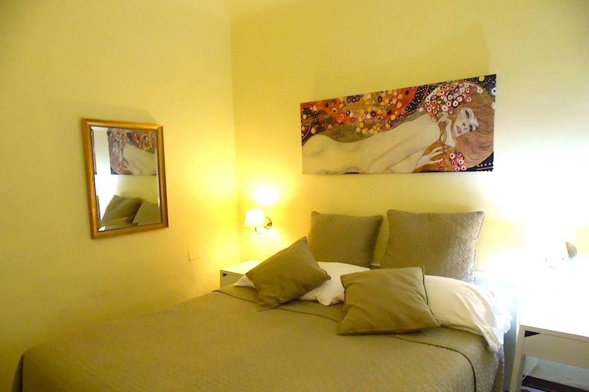 A bedroom with a bed, pillows, a nightstand, a lamp, a mirror, and a painting on the wall.