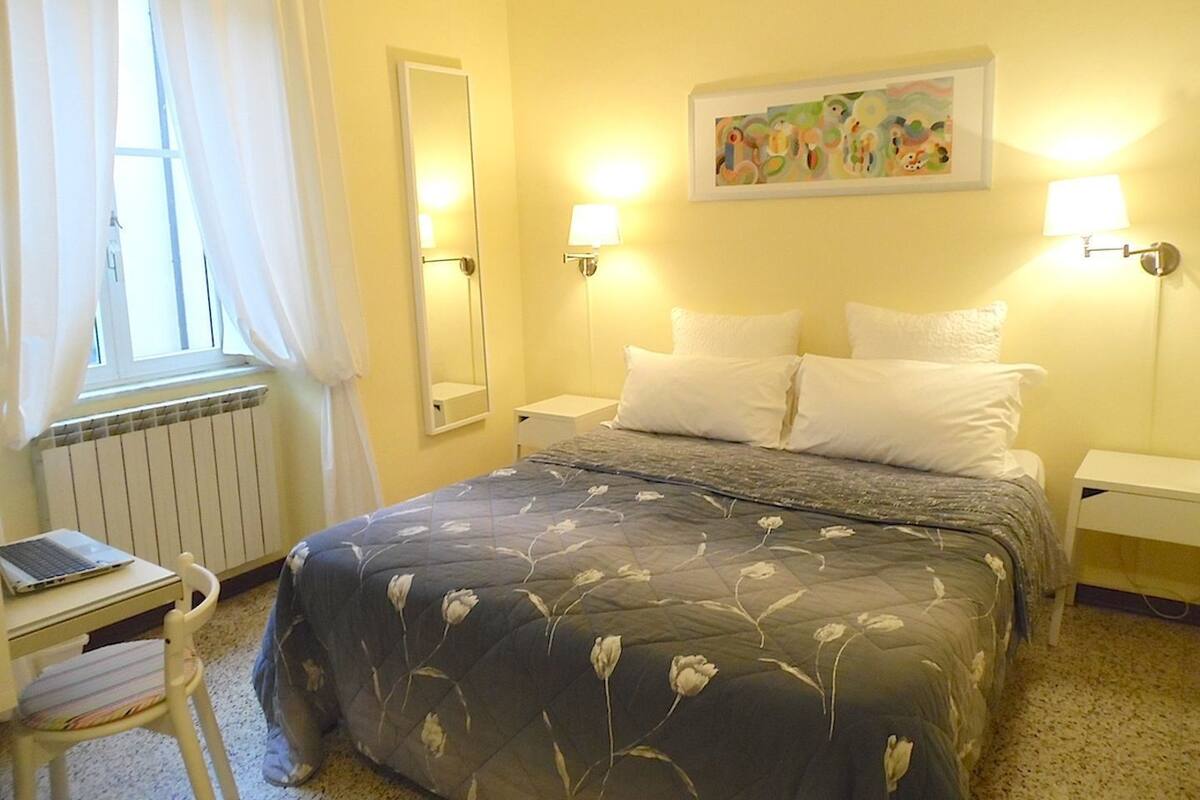 A hotel room with a bed, two bedside lamps, a desk with a laptop, a chair, a radiator, and a painting on the wall.