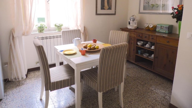 A dining area with a table set for two, chairs, a radiator, a window with curtains, a framed painting, a radio, and a cabinet with dishes. - B&B Casa Alba (Lucca)